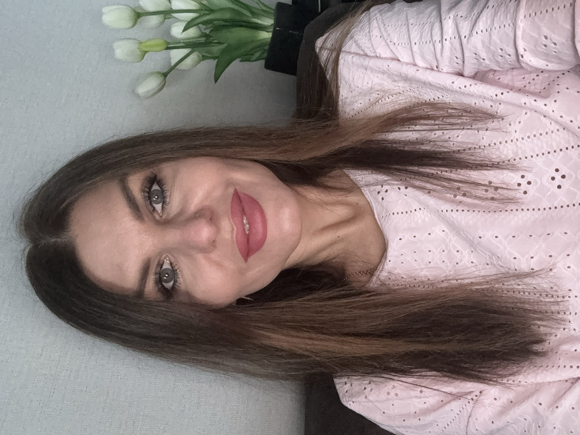A mid-angle, portrait-style photograph features a woman with light skin, light grey eyes, and brown hair with highlighted tips. She is wearing a light pink, eyelet-patterned top. The woman is smiling, displaying red lipstick. Behind the woman, to the upper left corner of the frame, are several white tulips in a dark vase. The background is a light grey wall on the left, and a dark brown or black object, possibly a piece of furniture, on the right. The location in Chaikovsky, Russia, is not deducible from this image.