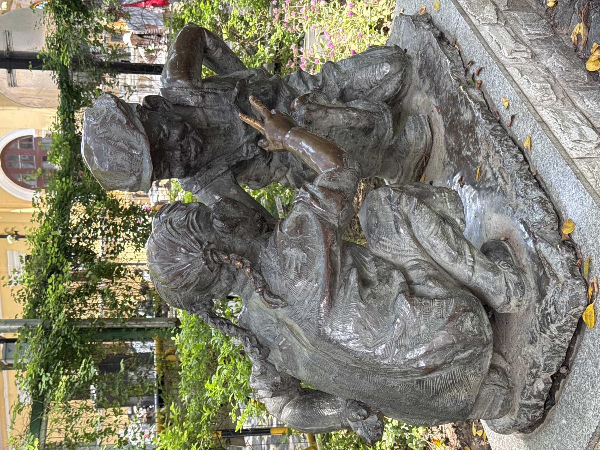 This image shows a bronze sculpture of two people located in the Liwan District, China. The sculpture depicts a girl with braided pigtails seated with legs crossed, and a man wearing a hat leaning over her, holding up his hand with two fingers extended. The sculptures are placed atop a grey stone base. Behind the sculpture are green plants and in the background, buildings typical of the area can be seen.