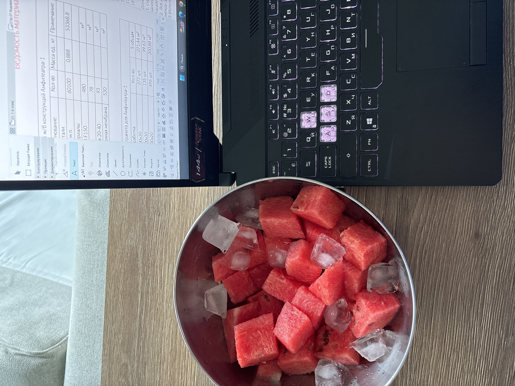The image shows a laptop, a bowl of watermelon with ice cubes, and a portion of a couch. The laptop screen displays a technical document in Russian, possibly related to architectural design or construction, with numerical data and diagrams. The keyboard of the laptop is visible, with some keys highlighted with a purple hue. A stainless-steel bowl filled with cubed watermelon pieces and ice is placed in the foreground. The table the objects are on is made of wood. A portion of a couch with beige fabric is visible in the background on the left side. There is no information in the provided images about the location of this scene in Choeng Thale, Thailand. There are no people present in the image.