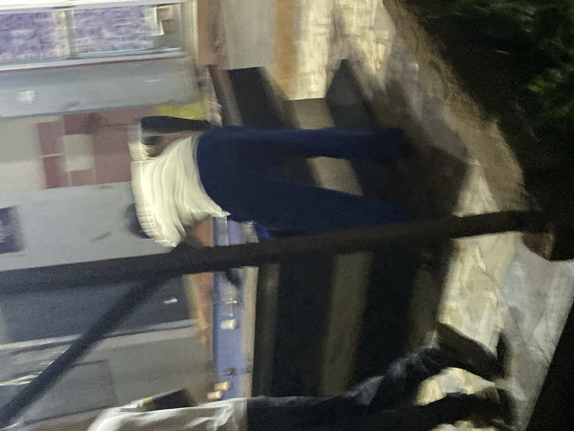 In Du, Nigeria, a person is captured in a blurred photograph, engaged in an activity that appears to be related to billiards or a similar table game. The individual, dressed in a light-colored t-shirt and dark pants, is positioned behind a table with a blue surface, possibly a billiard table, and is bent over as if to strike a ball. The background is indistinct, showing a building with windows and walls, suggesting an indoor or covered outdoor setting. The lighting is dim, contributing to the overall blurriness of the image, with some light sources visible in the distance, possibly from streetlights or interior lights. The ground appears to be paved or tiled. Another person, partially visible on the left side of the frame, is also dressed in dark clothing and is standing near the table. The scene suggests a casual recreational environment within the city.