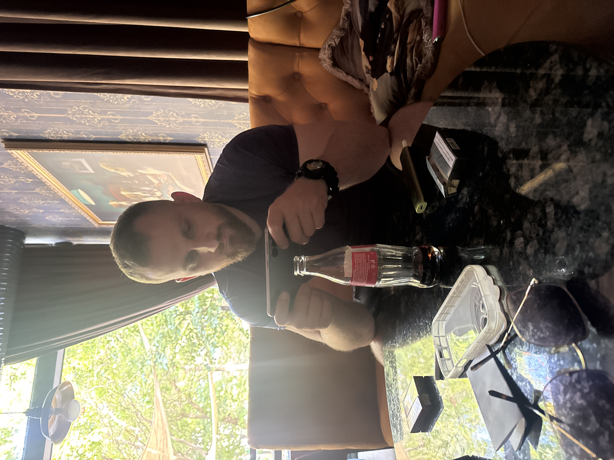 A man is sitting at a table in a dimly lit establishment. He is holding a smartphone horizontally and appears to be playing a game. A bottle of Coca-Cola, a pack of cigarettes, a phone case, a pair of sunglasses, and a vape pen are also on the table. The background shows a tufted orange sofa, patterned wallpaper, and a painting on the wall. Through a window, greenery and natural light are visible, suggesting the establishment might be near an outdoor area or garden. The overall ambiance suggests a relaxed, casual setting. The city of Varna, Bulgaria, is mentioned in the prompt, but there are no specific visual cues within the image to confirm this location.