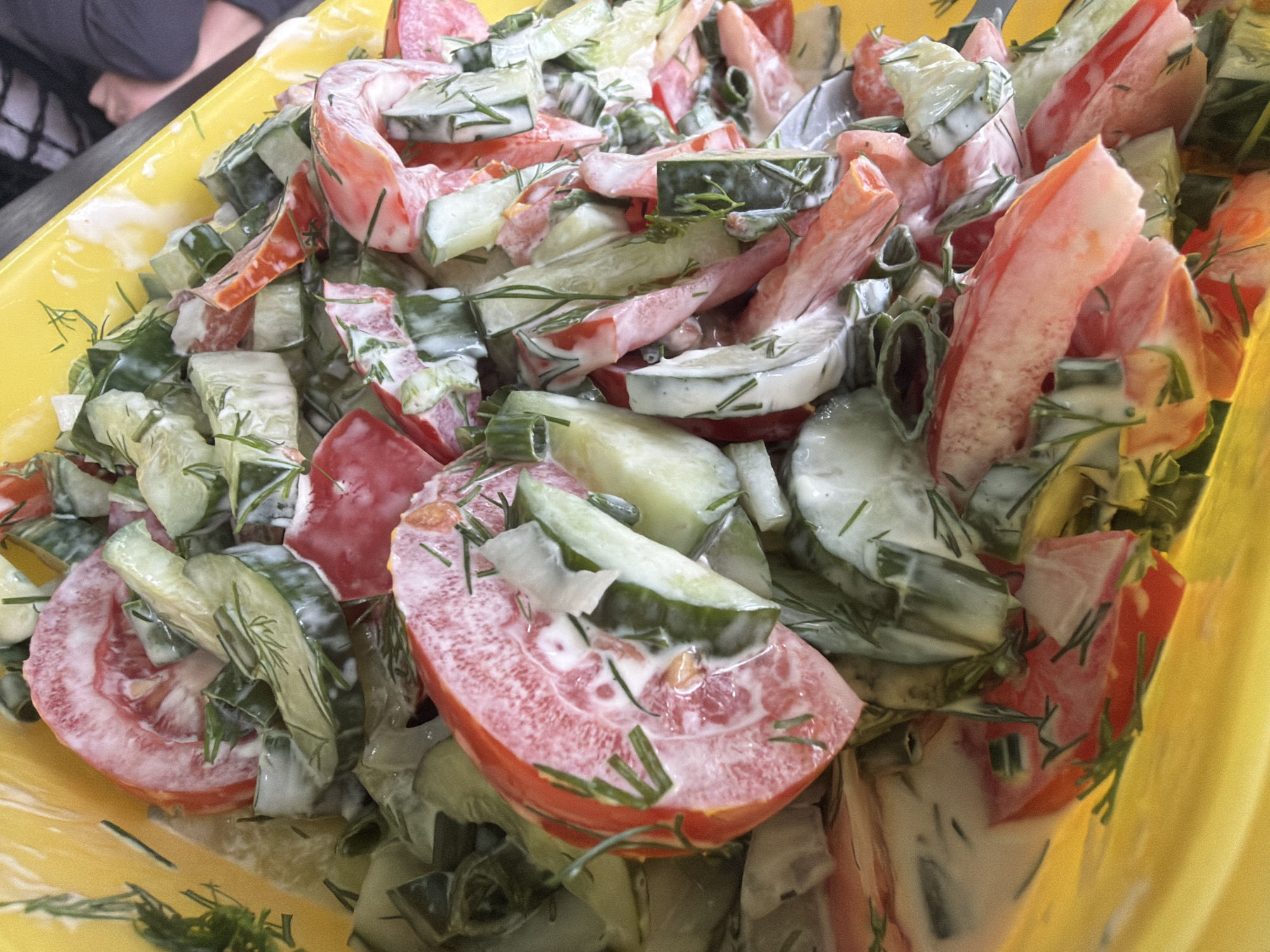 A close-up overhead shot shows a yellow bowl overflowing with a salad made of chopped tomatoes and cucumbers. The salad is dressed in a creamy white sauce and sprinkled with chopped dill. The background is blurred, but suggests an indoor setting, possibly a kitchen or dining area. No people are clearly visible in the image, though a person's arm and shoulder are visible in the upper right corner. The image does not provide any information about its location within the city of Petukhi, Russia, or any other location.