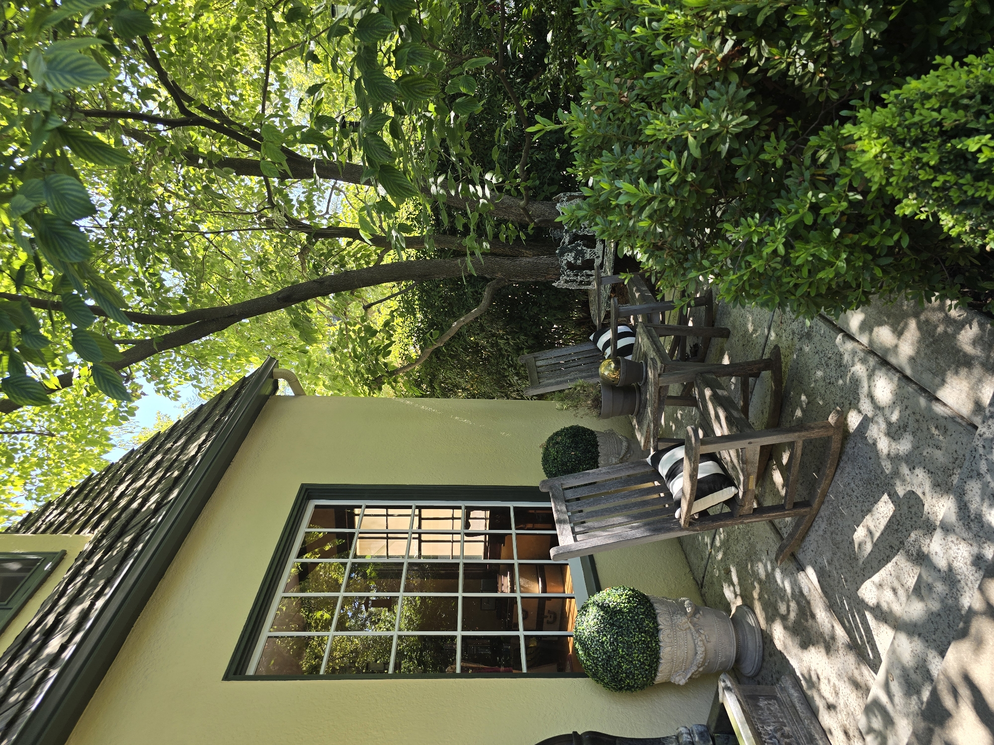 An outdoor patio area adjacent to a light yellow stucco house features two weathered wooden rocking chairs with black and white striped cushions. A small wooden table sits between the chairs. In front of the house is a large, decorative planter holding a spherical topiary. To the right, dense green bushes form a natural backdrop. Sunlight filters through the leaves of overhead trees, casting dappled shadows on the concrete patio. The scene is set in a residential area, possibly a garden or backyard.