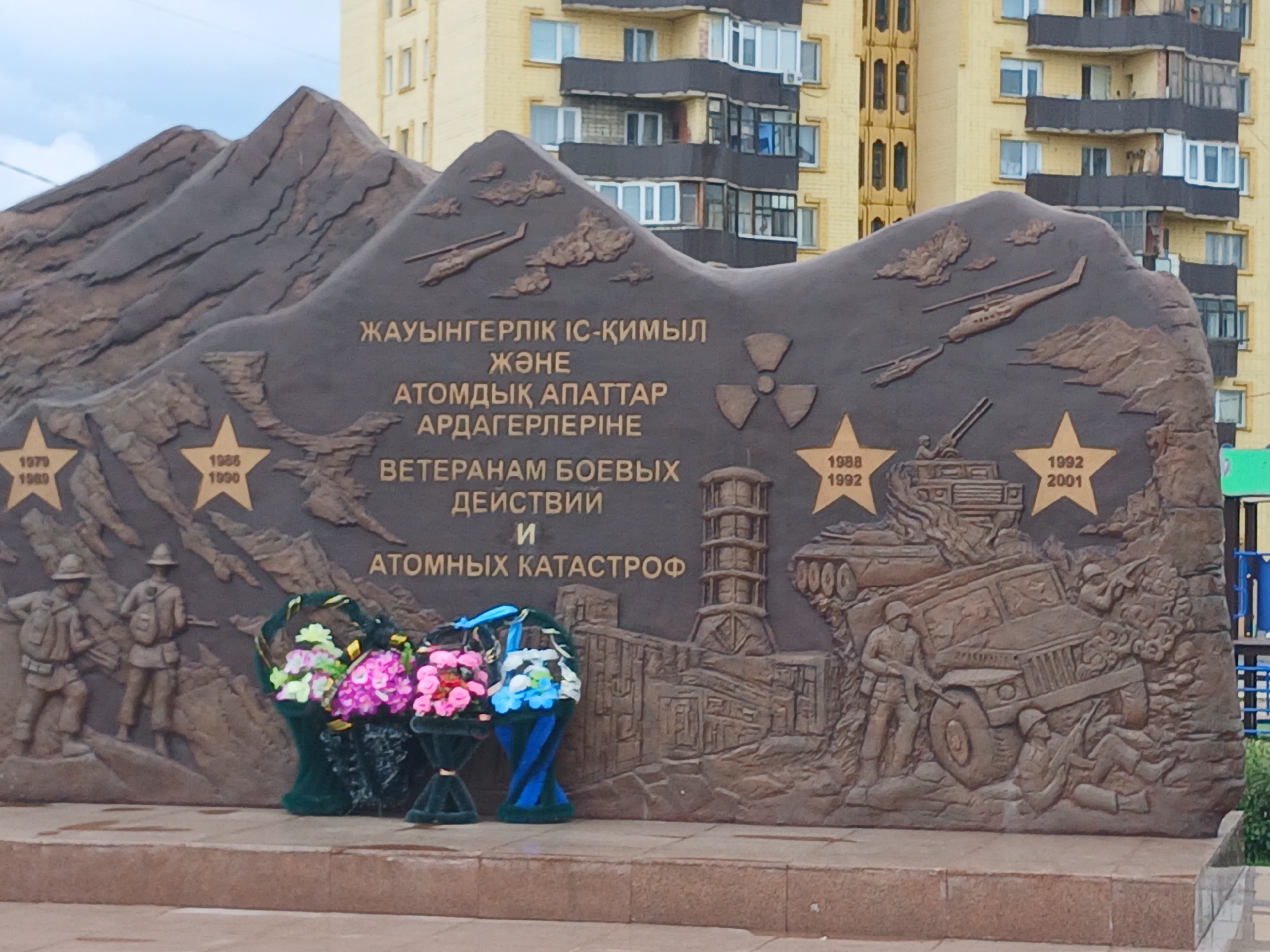 This is a close-up, eye-level, outdoor, daytime, medium shot of a large, relief sculpture in Shchuchinsk, Kazakhstan. The sculpture depicts mountainous terrain with various scenes. In the foreground, on the left, there are two figures of soldiers in combat gear with rifles. To the right of the soldiers is a large, stylized representation of a mushroom cloud and a nuclear symbol. Further to the right, a tall, cylindrical structure resembling a cooling tower is depicted, followed by a scene with a military vehicle, possibly a jeep, overturned with two soldiers in combat positions around it. The sculpture is made of dark-colored metal and is mounted on a stone base. Two large, green-covered wreaths with colorful artificial flowers are placed in front of the sculpture on the stone base. In the background, a multi-story apartment building with a yellow facade and dark balconies is visible. The sky is overcast. The sculpture appears to be a war memorial, commemorating military service and nuclear disasters.