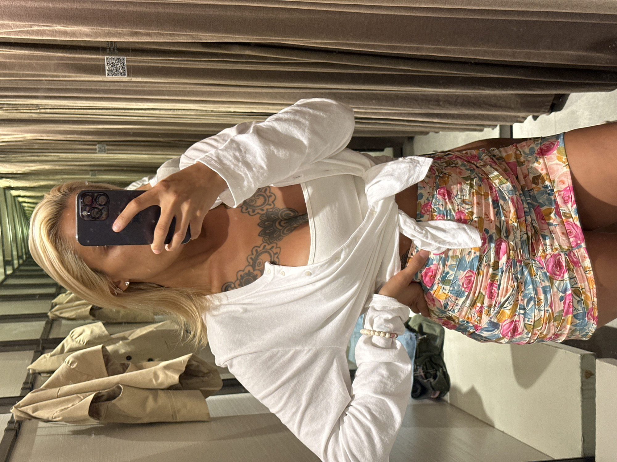 A person is taking a selfie in a fitting room. They are wearing a white long-sleeved shirt tied at the waist over a floral-print skirt. The person has blonde hair, tattoos on their chest, and is holding a smartphone to take the photo. The fitting room has beige curtains in the background and mirrors that reflect clothes hanging on racks. The image appears to have been taken indoors, likely in a clothing store. The lighting is artificial, suggesting it could be any time of day. No specific emotions are clearly discernible from the image, but the act of taking a selfie in a fitting room might suggest an interest in fashion or trying on new clothes. The location context is Kuta, Indonesia. There is a QR code visible on the wall in the background, which may be part of the store's branding or information system.