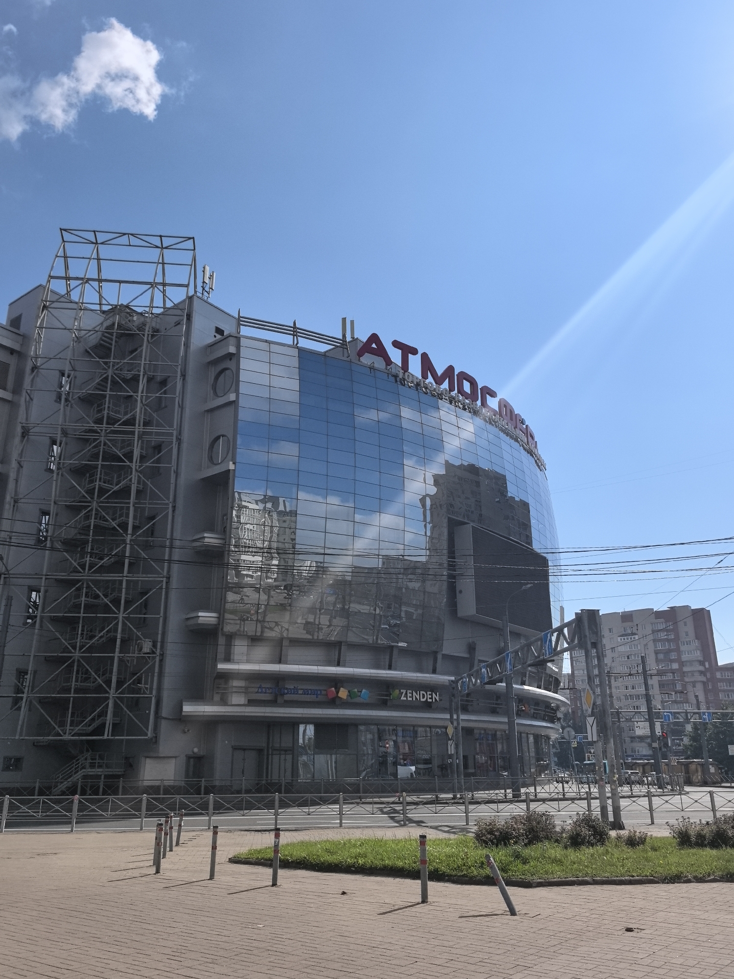 The image shows the "Atmosphere" shopping center located in the Moskovsky District of Saint Petersburg, Russia. The shopping center is a large, modern building with a distinctive curved facade covered in reflective glass. The building features a prominent red sign with the name "Atmosphere" at the top. To the left of the main building, there is an external metal staircase structure. The foreground shows a paved pedestrian area with bollards and a grassy strip. In the background, other buildings and power lines are visible under a clear blue sky with some clouds. The scene appears to be captured on a sunny day.