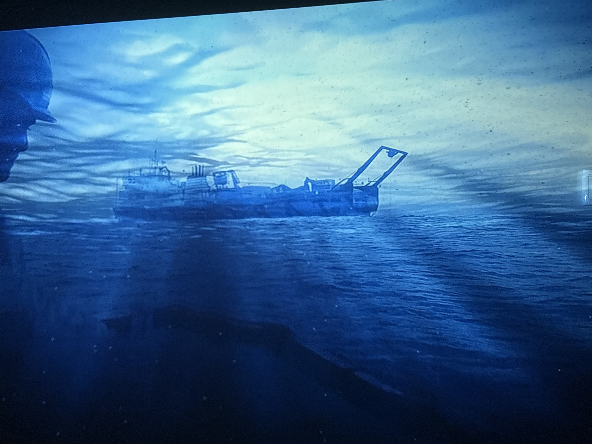 A blue-toned image depicts a ship on the water under a cloudy sky. The ship appears to be an older model, possibly a fishing vessel, with visible machinery and a crane-like structure at the stern. The water is textured with visible waves, and the light suggests it could be either dawn or dusk. A partial figure of a person is seen in the lower left corner. The location is unknown.