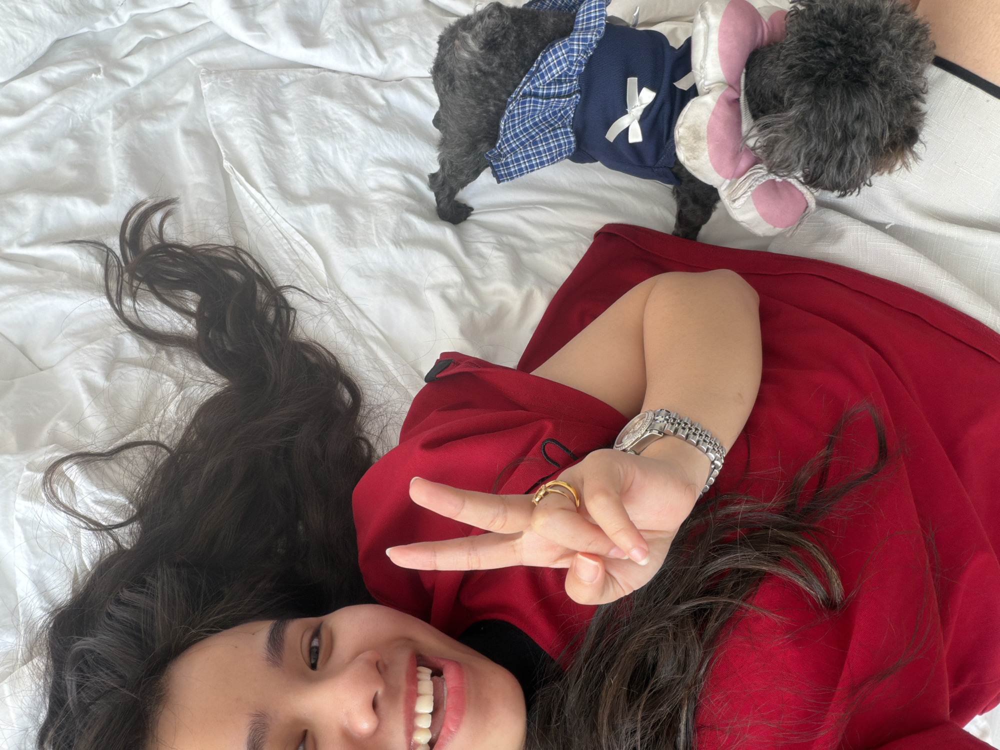A woman with long dark hair is lying on a white bed, smiling and giving a peace sign. She is wearing a red dress and a silver watch, and a gold ring is visible on her finger. Behind her, a small black dog wearing a blue and white checkered outfit and a stuffed animal is standing on the bed. The image appears to be taken from a top-down perspective. There is no visible evidence to support the image's location in the city of Quy Nhon Ward, Vietnam.