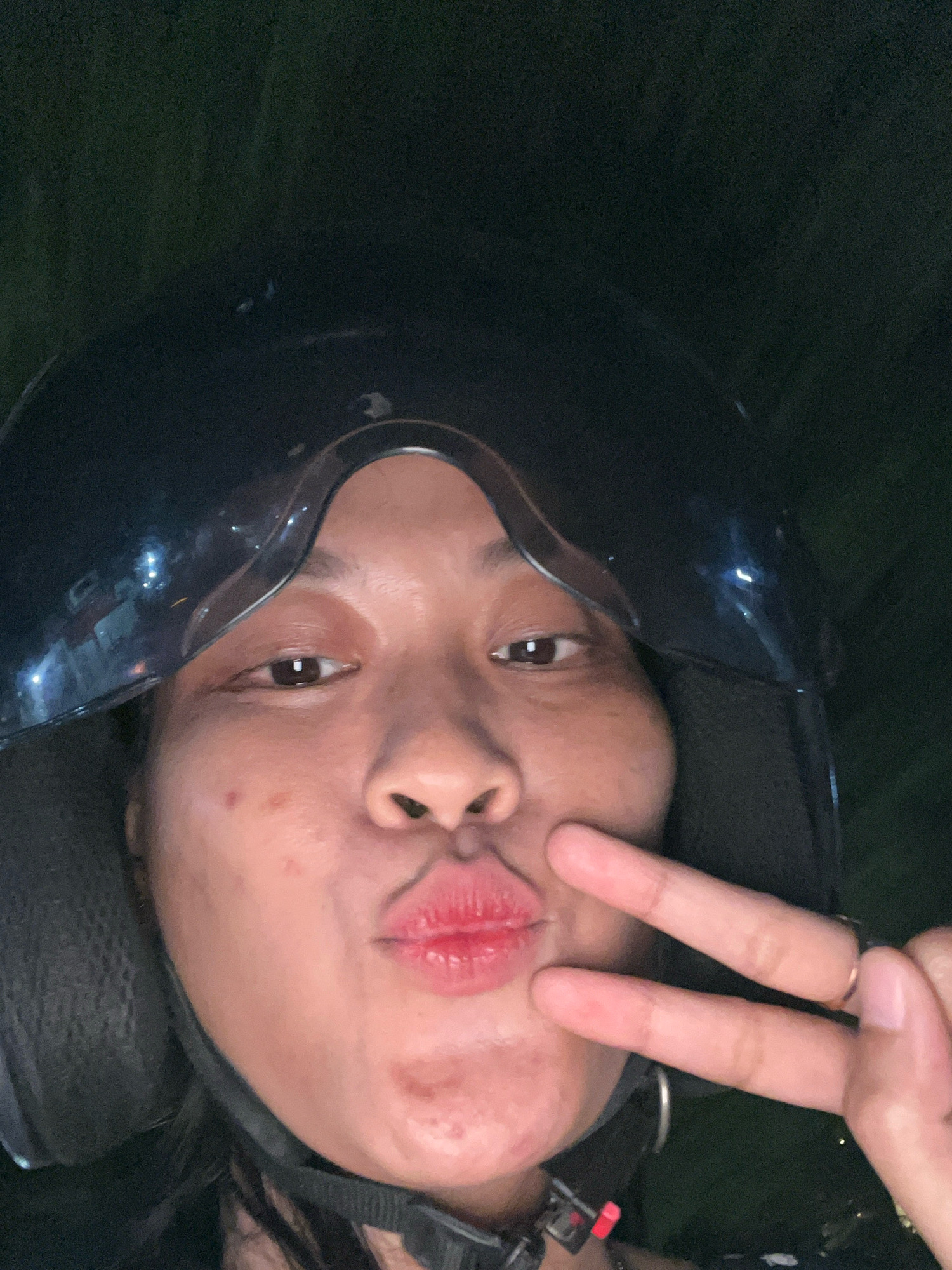 A close-up selfie captures a person wearing a black helmet, posing with puckered lips and giving a peace sign with their right hand. The person's face is illuminated, revealing light skin with some minor blemishes. Their lips are colored with red lipstick. The background is dark and appears to be outdoors, possibly at night, with some blurred greenery or foliage visible. The helmet's reflective surface shows some faint light sources, suggesting artificial lighting in the vicinity. The image is taken at a low angle, looking up at the subject. No specific details about the location within Kecamatan Pati, Indonesia, can be ascertained from the image alone.