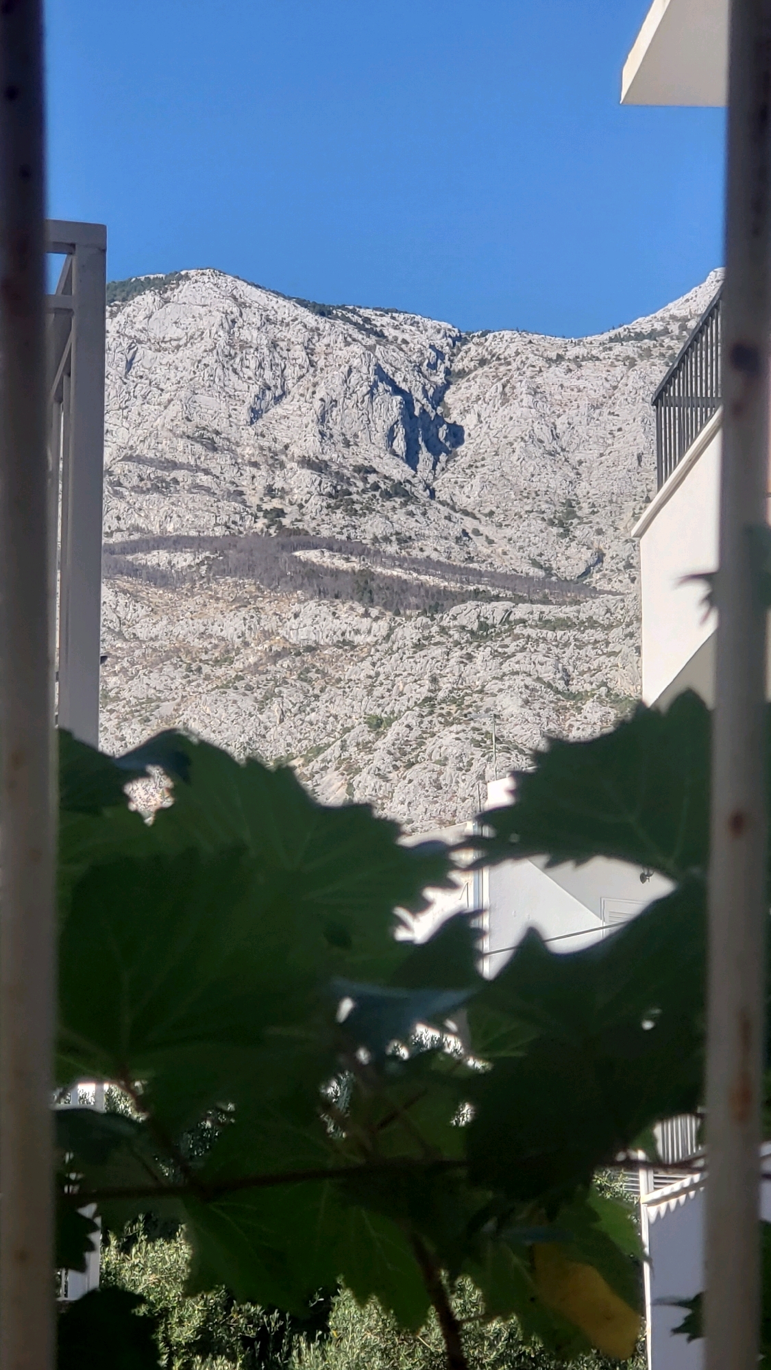 The image depicts a view of a rocky, mountainous terrain against a clear blue sky, framed by foreground foliage and parts of a building's exterior. The mountain range, characterized by its rugged, light-colored rock formations and sparse vegetation, dominates the background. A distinct, dark shadow casts a curved shape across the middle section of the mountain slope. In the mid-ground, sections of a white building with a black railing are visible, suggesting an urban or residential setting. The foreground is partially obscured by large, green leaves, some of which are backlit by the sun, creating a dappled effect. The overall scene suggests a landscape view from a balcony or window, possibly in a coastal or mountainous region.
This image was captured from Općina Tučepi, Croatia, a municipality on the Dalmatian coast known for its scenic beauty and proximity to the Biokovo mountain range. The rocky slopes and clear blue sky are characteristic of the Mediterranean climate found in this region. The presence of the building indicates that the viewpoint is from a structure overlooking the natural landscape.