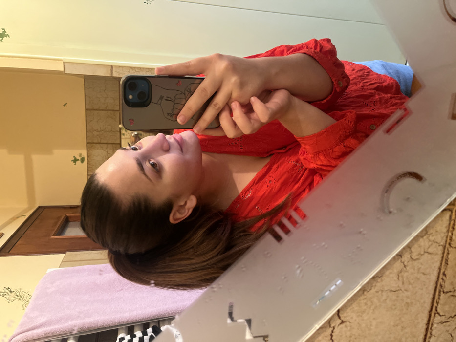A female individual with dark brown hair, styled in a high ponytail, is depicted taking a mirror selfie in what appears to be a bathroom in Ancenis, France. She is wearing a red, long-sleeved, eyelet-detail blouse and light blue bottoms. Her right arm is raised, holding a dark-cased smartphone with a line drawing of hands and a red heart sticker on the back. Her reflection captures her in profile, gazing towards the phone. The mirror features a frosted decorative border with geometric cutouts and water droplets. The background, reflected in the mirror, shows light-colored walls adorned with small green frog decals, a section of brown textured wall tiles, a light purple towel hanging on a rack, and a dark wooden door in the upper left quadrant. A light-colored vertical panel or door frame is visible on the far right of the reflection.
