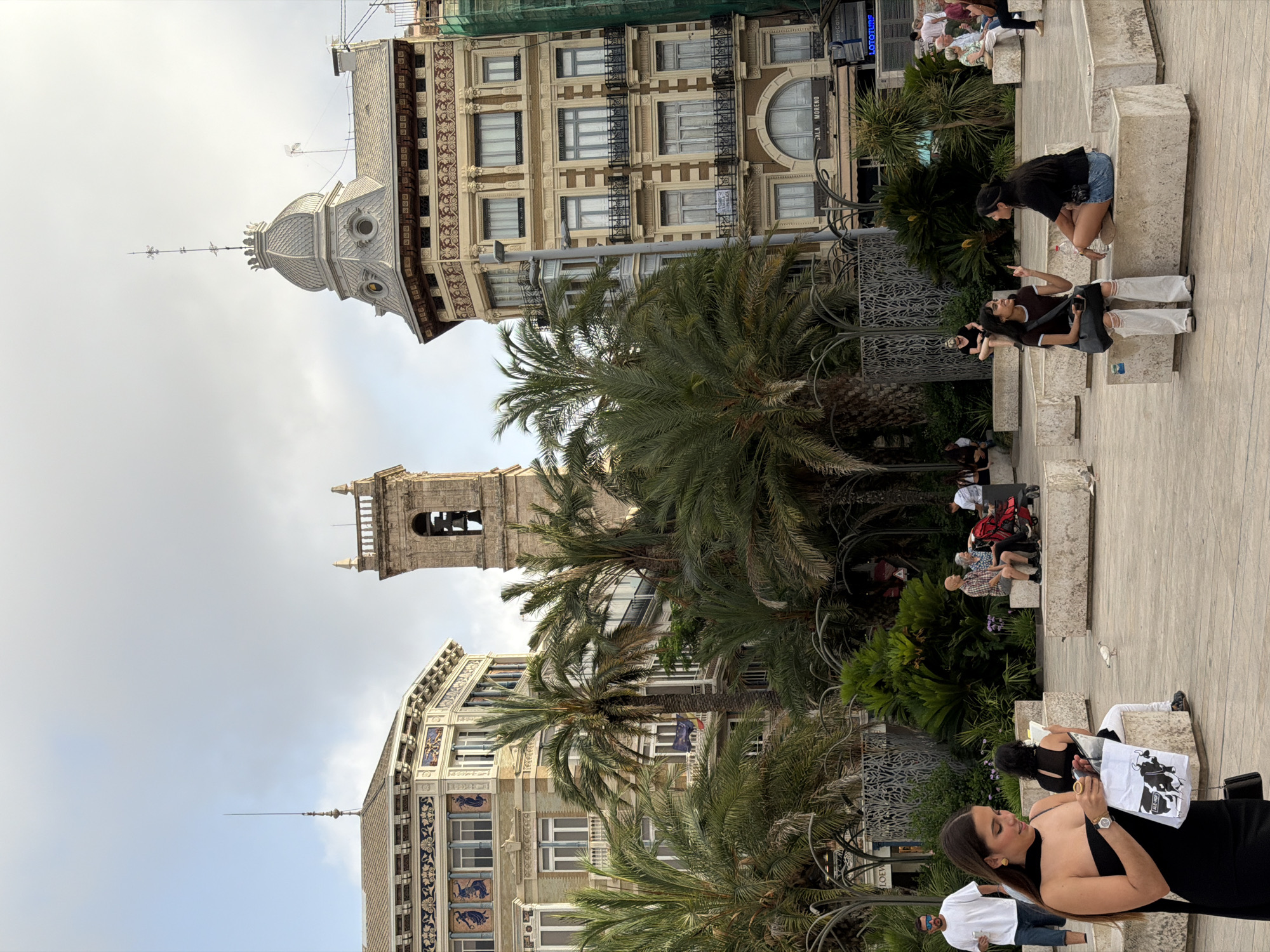 In an outdoor urban square, two young women sit on concrete benches facing each other, engaged in conversation. One woman, on the left, wears a dark top and light-colored ripped jeans, with a dark crossbody bag. The other woman, on the right, sports a black t-shirt and denim shorts, with a dark bag slung over her shoulder. A water bottle rests between them. In the foreground, a woman in a black strapless dress walks away from the camera, holding a white paper bag with cow illustrations and text that reads "ALE-HOP". Other people are visible in the background, including a man in a white t-shirt and shorts, and a couple with a baby stroller, seated on a bench. Several palm trees are scattered throughout the scene, and prominent architectural structures, including a bell tower and ornate buildings, frame the background. The sky is overcast.