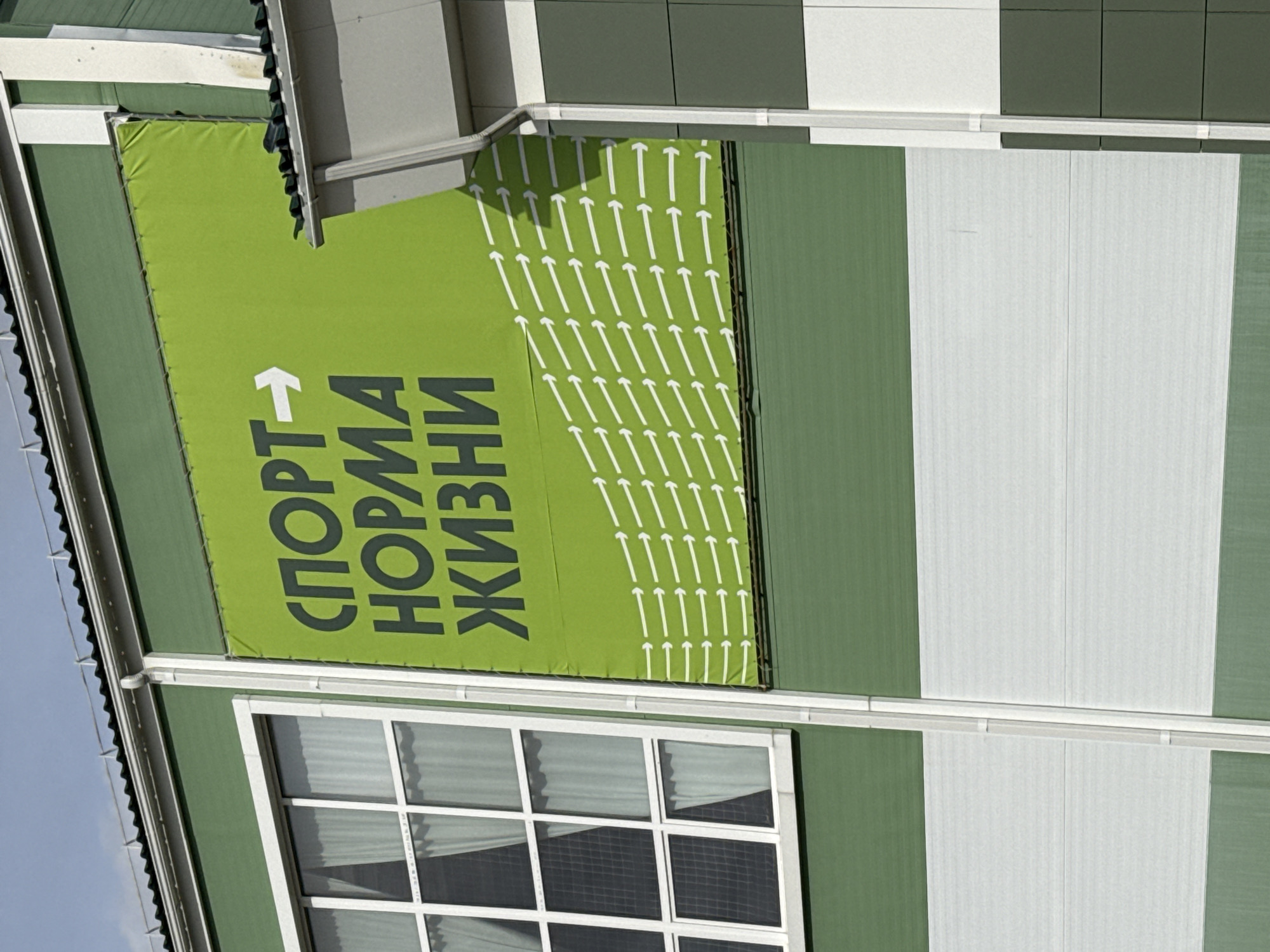 The image shows a building with a green and white exterior. A green banner with white arrows and Russian text is mounted on the building. The text reads "СПОРТ-НОРМА ЖИЗНИ" (Sport is the norm of life). The building has a window with a white frame. The photo was taken in Tobolsk, Russia.
