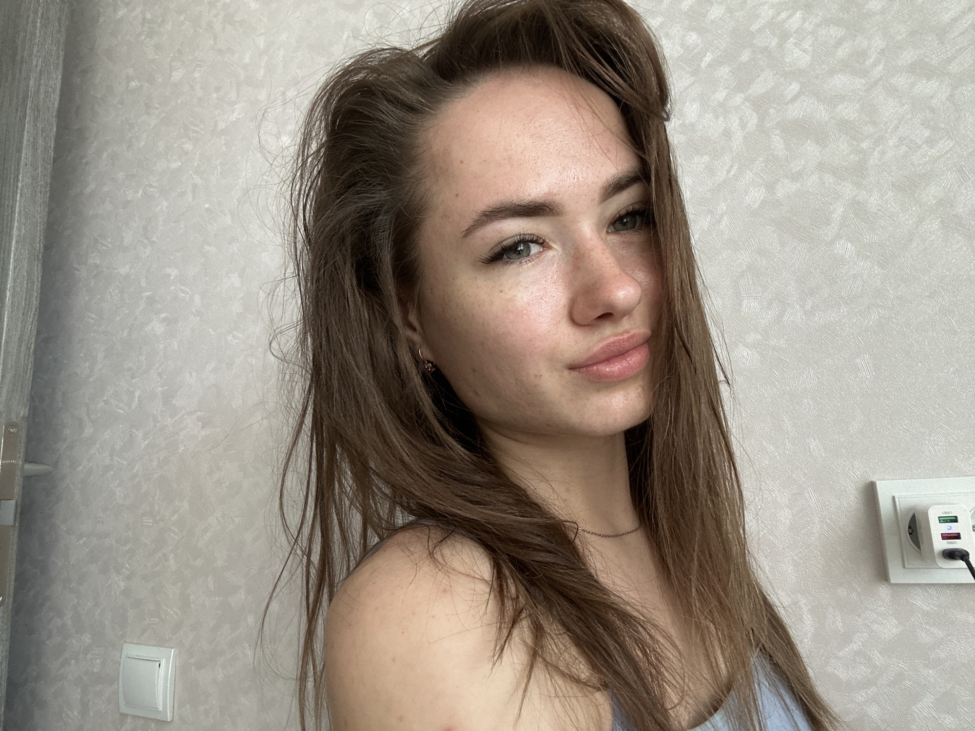 A young woman with long, medium-brown hair and light-colored eyes is captured in a waist-up portrait, looking directly at the camera with a subtle smile. She has noticeable freckles across her nose and cheeks and is wearing a light blue or gray top. A thin silver chain necklace adorns her neck, and a small dark earring is visible on her left earlobe. Her hair, which appears slightly disheveled, frames her face and extends over her left shoulder.
The background features a light-colored, textured wallpaper. On the far left, a vertical grey element, possibly a door frame or the edge of a door, is visible, along with a white light switch mounted on the wall below it. On the right side of the frame, a white electrical outlet contains a dual USB wall charger. The charger has "USB1" and "USB2" printed next to its ports, which feature green and red/purple accents, and "c-sinn" is visible on its lower body. The lighting is bright and even, suggesting a daytime indoor setting.