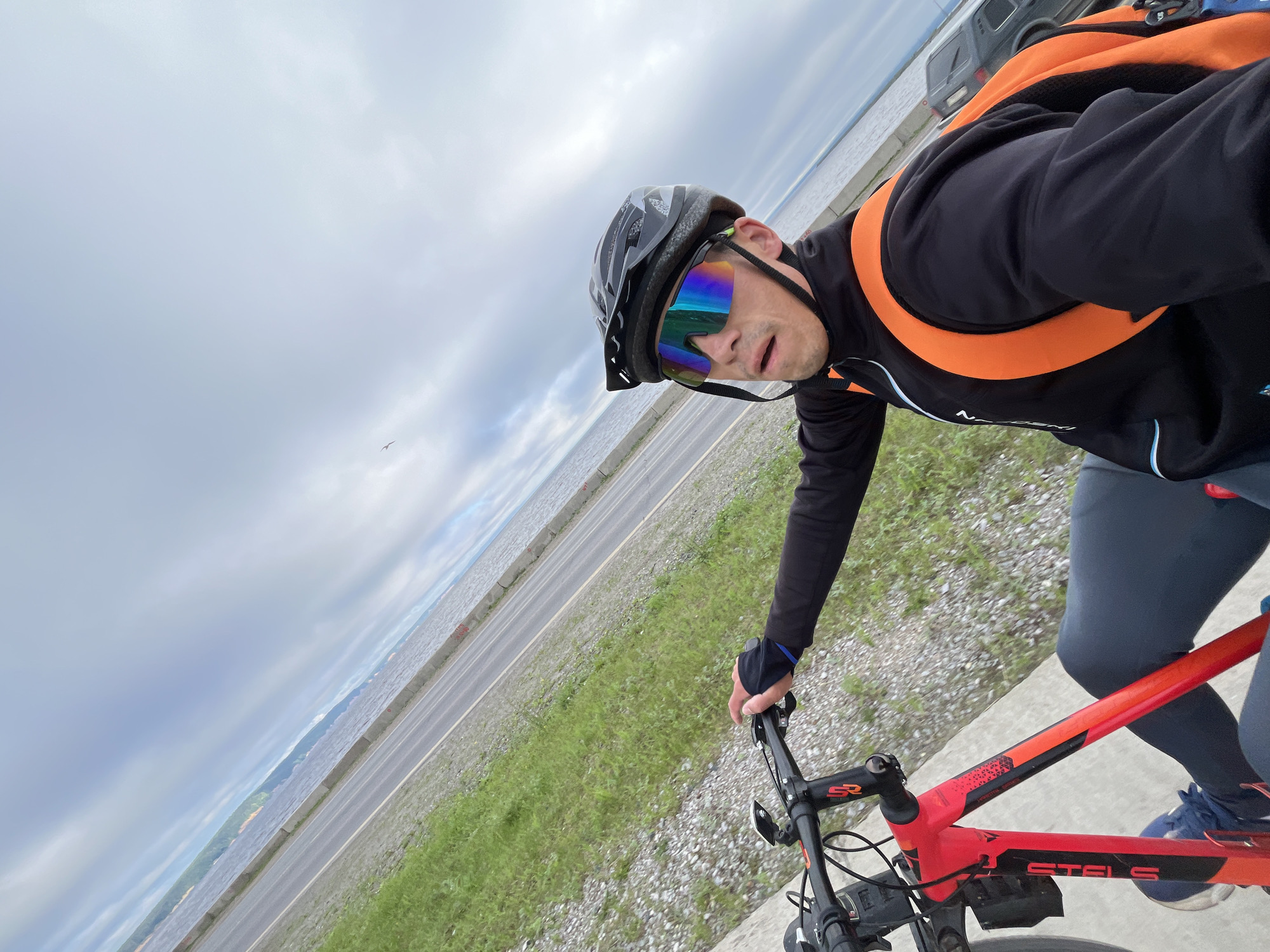 In a selfie taken from a first-person perspective, a person is riding a red bicycle on a paved path next to a body of water. The cyclist is wearing a black helmet, large mirrored sunglasses, a black long-sleeved shirt with an orange backpack, and dark leggings. The path is bordered by grass and gravel. In the background, a wide river or lake is visible, with a distant shoreline and a cloudy sky. A dark SUV is parked on the road to the right of the cyclist. The scene appears to be along the embankment of the Kama River in Chaikovsky, Russia.