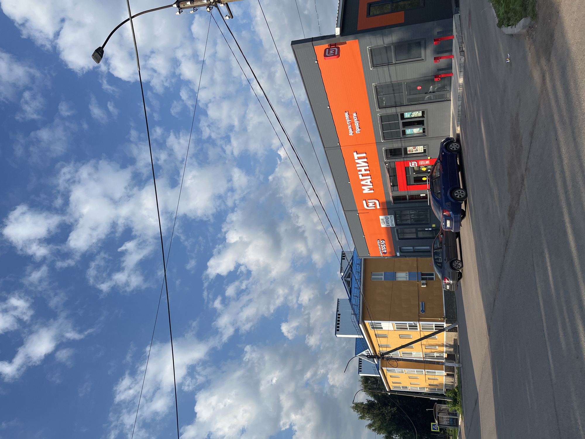 The image captures a street view in the city of Blagoveshchensk, Russia, under a bright blue sky with scattered white clouds. A modern commercial building with an orange and grey facade dominates the right side of the frame. The building features large windows and prominent signage for "МАГНИТ," a Russian supermarket chain, and also advertises "Аптека" (pharmacy) and "Продукты" (groceries). Two cars are parked in front of the building: a dark blue sedan on the right and a grey sedan to its left. To the left of the commercial building, a multi-story residential building painted in yellow with blue roofing and white window frames is visible. In the background, trees and a bus stop can be seen. Electrical wires crisscross the sky, supported by a utility pole on the far right. The street in the foreground is paved, with shadows cast by an unseen light source.