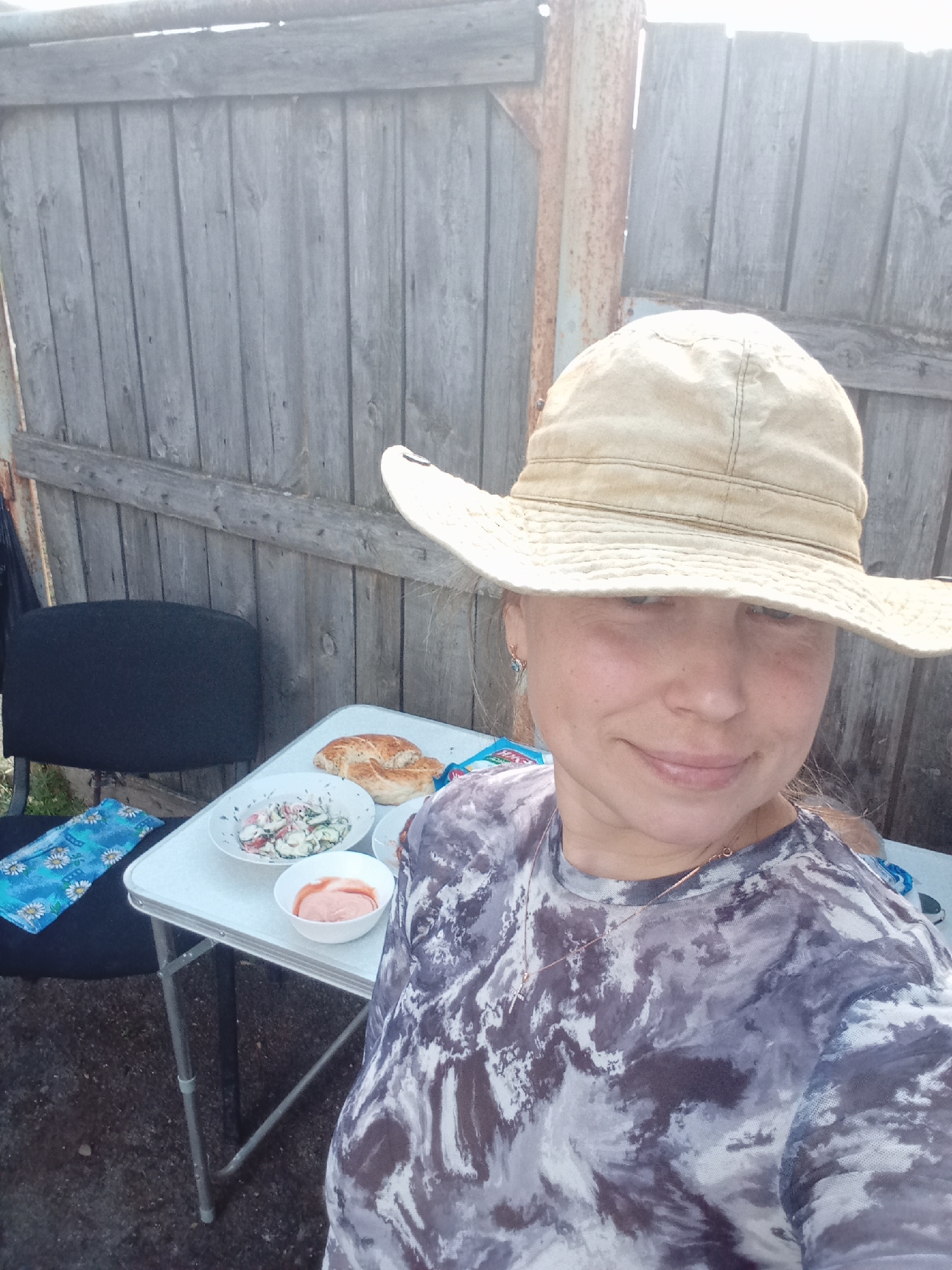 A woman wearing a tan hat and a gray tie-dye shirt stands in front of a weathered gray wooden fence. A white folding table is positioned next to her, displaying a plate of salad, a dish of pink sauce, and a piece of bread. A black office chair with a blue floral cushion sits behind the table. The location appears to be outdoors. The city of Кодинка, Russia is not discernible from the image.