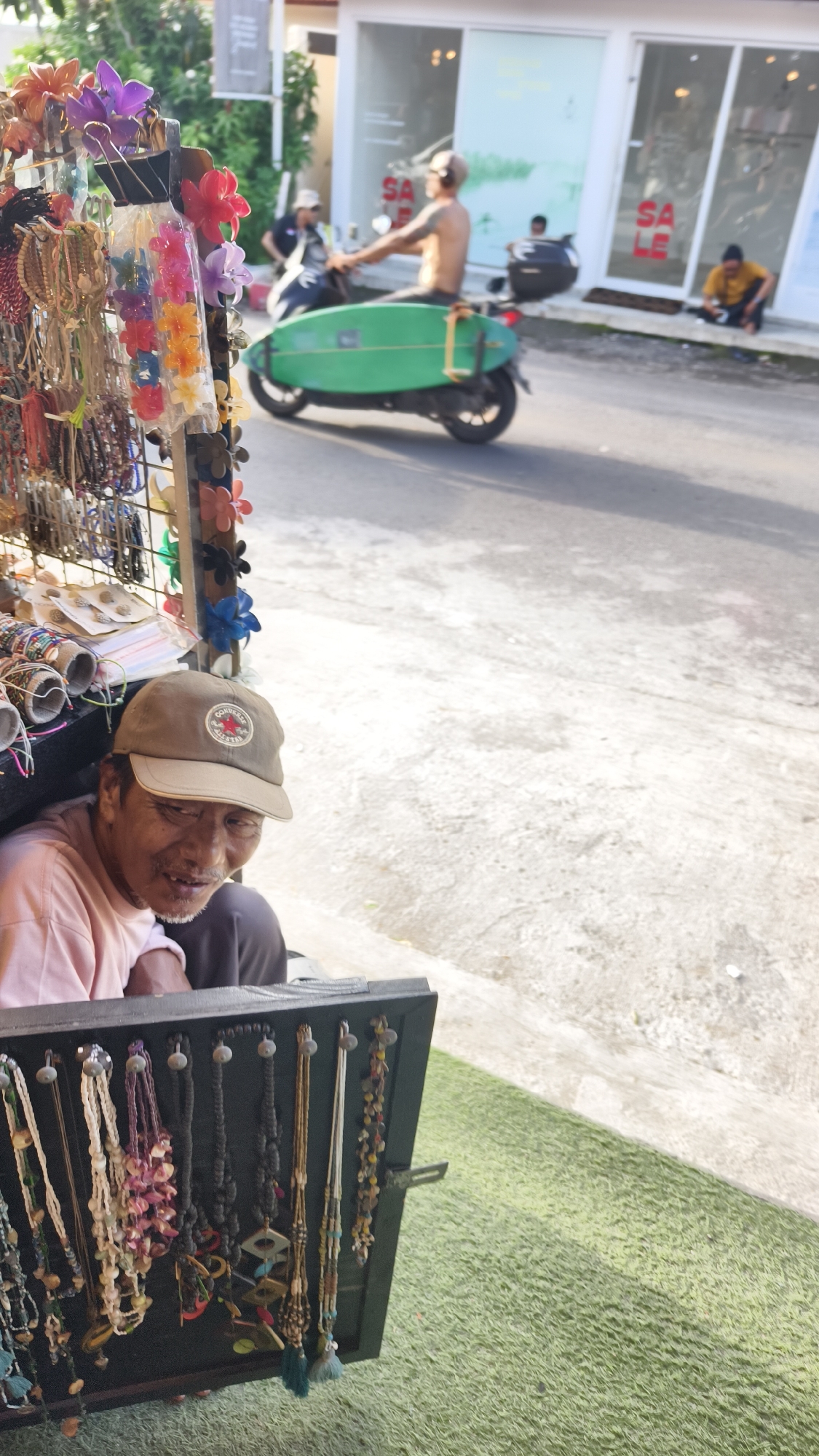 An outdoor scene in Pecatu, Indonesia features a vendor in the foreground positioned behind a display rack containing an assortment of jewelry and colorful hair accessories. The vendor, wearing a light pink shirt and a tan-colored baseball cap with a logo, is gazing forward with a slight smile.
In the blurred background, a shirtless man riding a scooter with a green surfboard strapped to the side is moving along the street. There is a white building with large glass windows that reflect the environment. A sign on the window displays the word "SALE" in bold, red letters. Another man is seated on the sidewalk near the building.
The ground is a mixture of asphalt and a patch of artificial green turf. The lighting suggests it's daytime, with shadows indicating strong sunlight. Other pedestrians and vehicles are visible in the far background, contributing to the street's atmosphere.