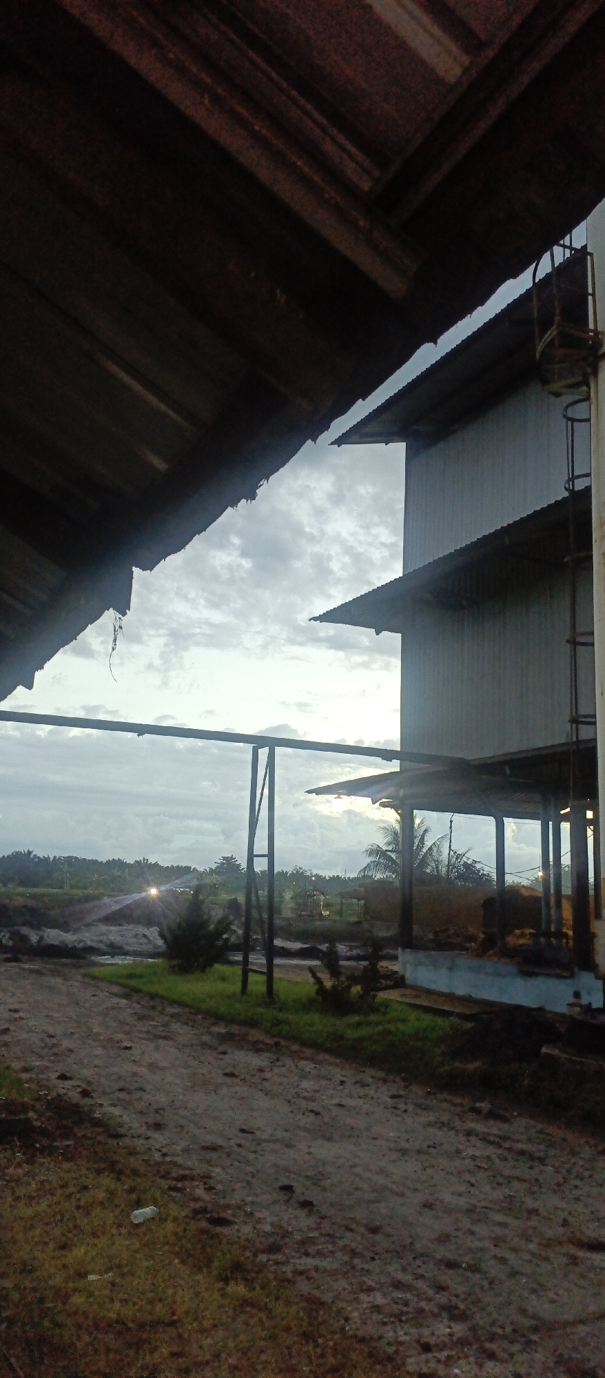 The image depicts an outdoor industrial or agricultural setting, likely near Iemirah, Indonesia. In the foreground, a dirt path is visible, with sparse green grass along its edges. To the left, the underside of a dark, corrugated metal roof is prominently featured, with rough, unfinished edges and visible structural beams. To the right, a multi-story building with corrugated metal siding rises, featuring several tiered, overhanging roofs. A metal ladder is attached to the side of this building.
In the middle ground, a metal frame structure stands with horizontal beams. Beyond this, a landscape of dense green foliage, possibly palm trees, stretches across the horizon under a cloudy sky. Two bright headlights are visible in the distance, suggesting vehicle movement or stationary lights. Some low-lying structures, possibly sheds or open-air shelters, are scattered within the foliage. The overall lighting suggests either dawn or dusk. There are no people visible in the image.