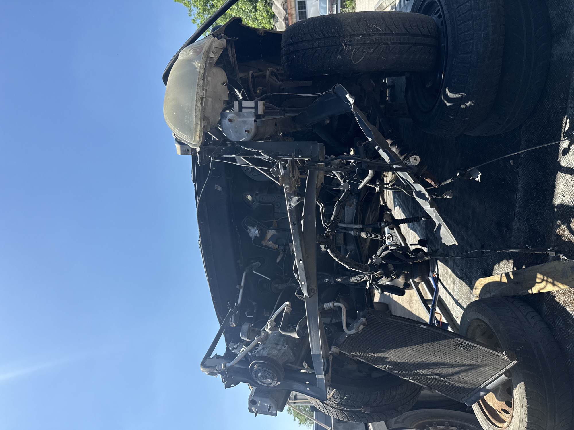 The image shows the front end of a dismantled car on what appears to be a trailer or flatbed. The car's engine compartment is exposed with various components such as the radiator, engine block, wiring, and headlight visible. Several tires are stacked on the trailer, positioned near the dismantled car. The sky is clear and blue, suggesting a daytime scene. While the image does not provide specific details for identifying the precise location in Shumen, Bulgaria, the setting implies a scrap yard, auto repair shop, or vehicle transportation context. No people are visible.