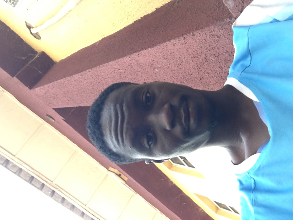 A male individual with dark skin, short black hair, a mustache, and a goatee is depicted in a close-up, upward-angled shot. He is wearing a light blue V-neck shirt with white trim. The subject looks directly at the camera with a neutral expression. The background features architectural elements: a textured reddish-brown wall prominently to the right, a light yellow wall with two white-framed windows to the left, and a beige or white eave structure overhead, indicating an outdoor setting adjacent to a building. This scene takes place in the city of Pindiga, Nigeria.