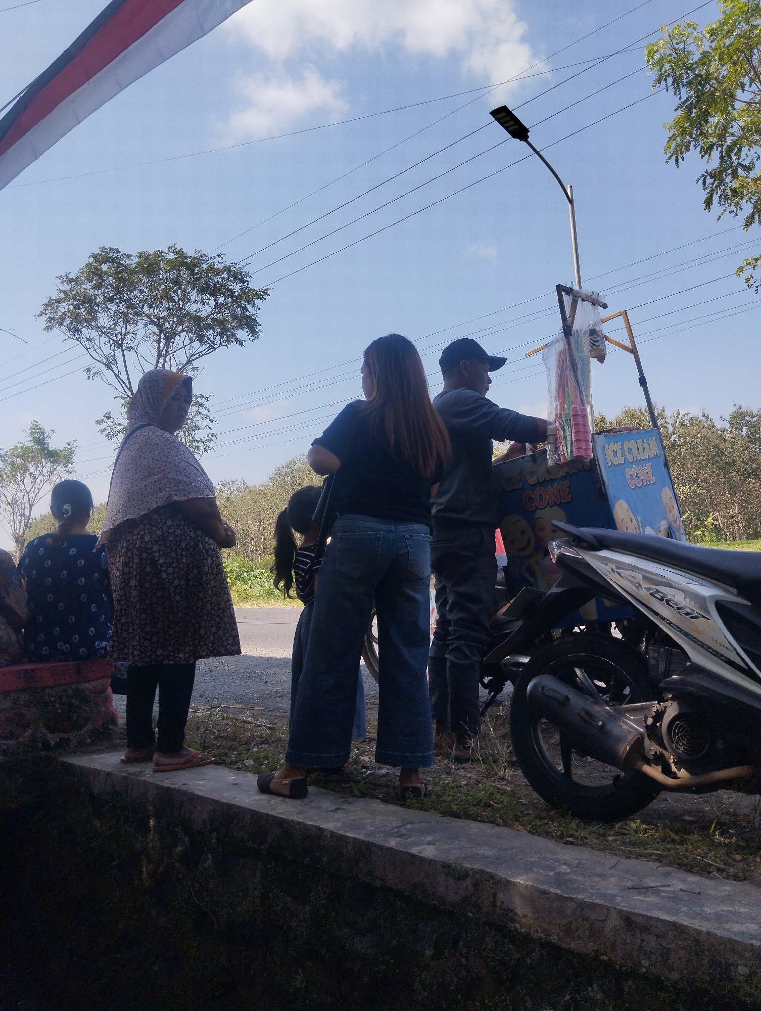 The image captures an outdoor scene on a bright, sunny day, possibly in Karangdoro, Indonesia, given the context of the provided information, depicting a roadside vendor. In the foreground, a concrete ledge runs horizontally, with a dark, possibly water-filled, ditch below it. On the ledge, several individuals are present. To the left, a person with dark hair pulled back sits on a red bench, facing away from the camera. Next to them, an older woman wearing a headscarf and a patterned dress stands, looking to the side. To the right of the woman, a young child with dark pigtails is partially visible. Further right, a woman in a black top and blue jeans stands with her back to the camera.
Dominating the mid-ground is an ice cream cart, supported by a wooden frame, with a light-colored pole extending upwards holding a solar-powered street lamp. The cart itself is blue with "ICE CREAM CONE" written in white text, and features images of cartoon faces. A vendor, wearing a dark long-sleeved shirt and a black baseball cap, is attending to the cart, appearing to serve customers. Several plastic cups and what seem to be ice cream cones are visible on the cart.
A motorbike is parked to the right of the ice cream cart, partially obscuring the lower portion of the cart and the vendor. The motorbike is white and black with visible branding.
The background shows a line of trees and vegetation, suggesting a rural or semi-rural setting. Overhead, power lines crisscross the bright blue sky, which has a few scattered white clouds. A red and white banner is visible in the upper left corner of the image. The overall impression is a snapshot of everyday life at a roadside stop.