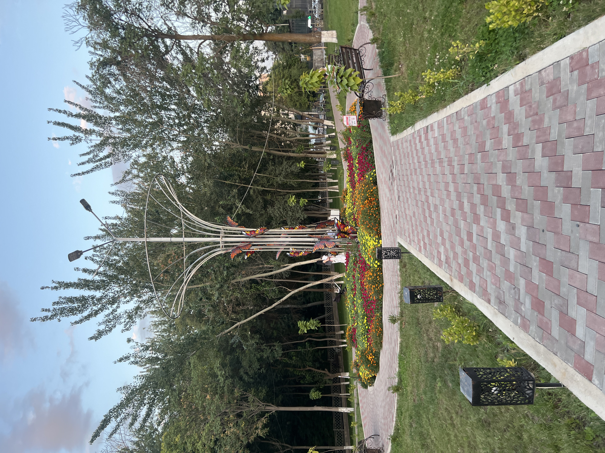 The image shows a park in Osh City, Kyrgyzstan, with a paved walkway made of red and gray bricks leading towards a central floral display. The flower bed is filled with a variety of colorful flowers, including red, orange, and yellow blooms. In the center of the flower bed stands a tall, metallic sculpture resembling a tree, adorned with decorative butterfly figures. Several trees with green foliage surround the park. Two benches are visible on either side of the walkway. In the background, a fence and more trees can be seen, along with a glimpse of a street with a few cars. The sky is partly cloudy.