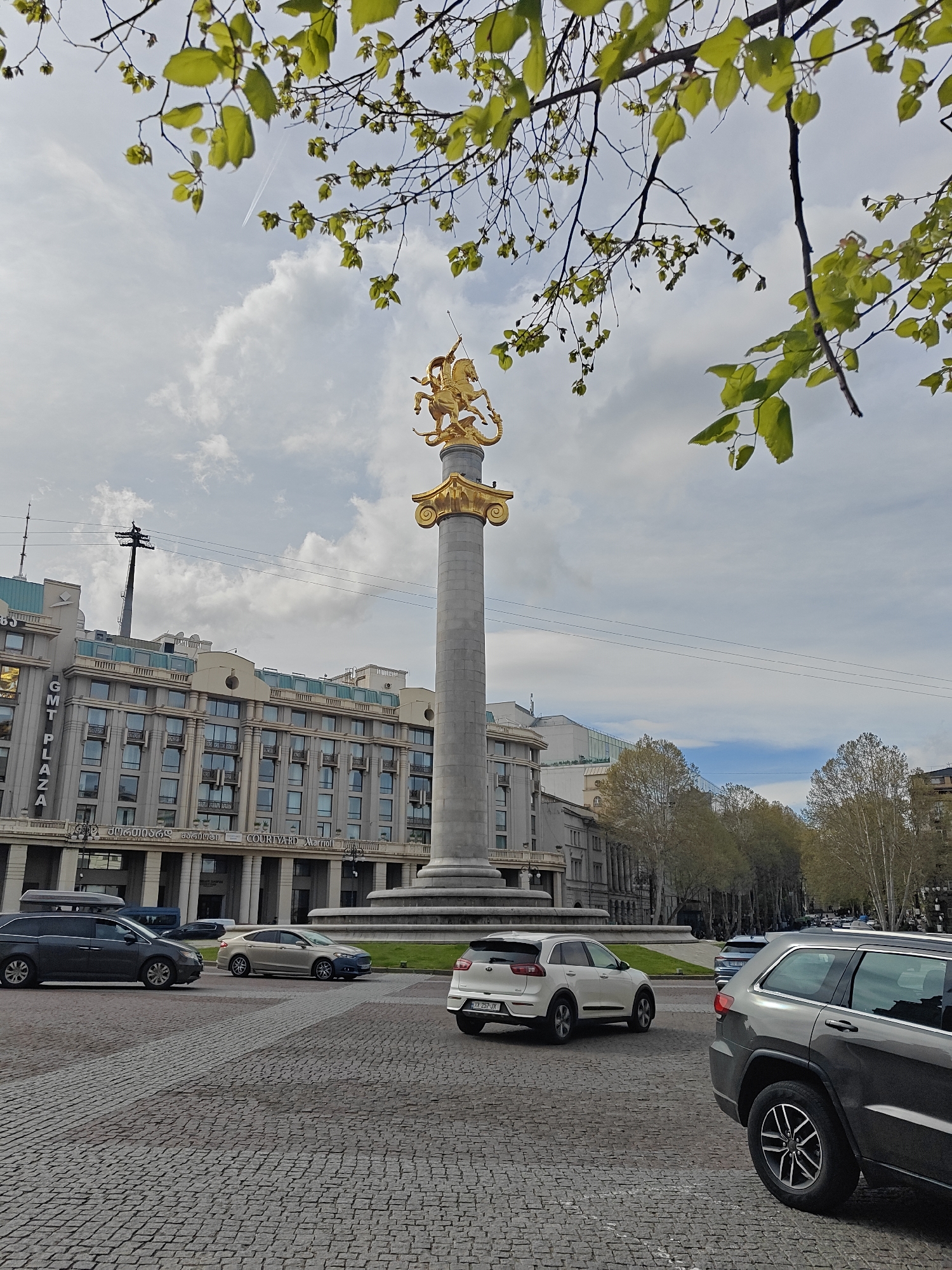 The image presents a vibrant urban square in Tbilisi, Georgia, under a bright, cloudy sky, indicative of daytime. Central to the scene is a majestic, tall grey column crowned with a striking golden equestrian statue, likely depicting Saint George slaying the dragon, a significant emblem for Georgia. This monument stands proudly on a circular, grassy base, surrounded by a wide expanse of cobblestone paving.
To the left, a large, modern building with a light-colored facade and numerous windows frames the square. Prominently displayed on its exterior are the texts "GMT PLAZA" vertically and "COURTYARD Marriott" horizontally at its base. Below "Courtyard Marriott", Georgian script is visible, which translates to "Hotel Tbilisi Marriott".
The square is active with several vehicles, including a dark grey SUV in the immediate foreground, a white SUV (with license plate "KX 257-JK"), a champagne-colored sedan, and a dark minivan, suggesting typical city traffic or parking. Overhead, power lines crisscross the sky, and a faint airplane contrail is visible against the clouds in the upper left. The top edge of the frame is adorned with fresh, bright green leaves on tree branches, clearly indicating the season as spring. In the background, other deciduous trees with sparser foliage line what appears to be a street or parkway. The overall scene captures the dynamic atmosphere of a central city landmark.