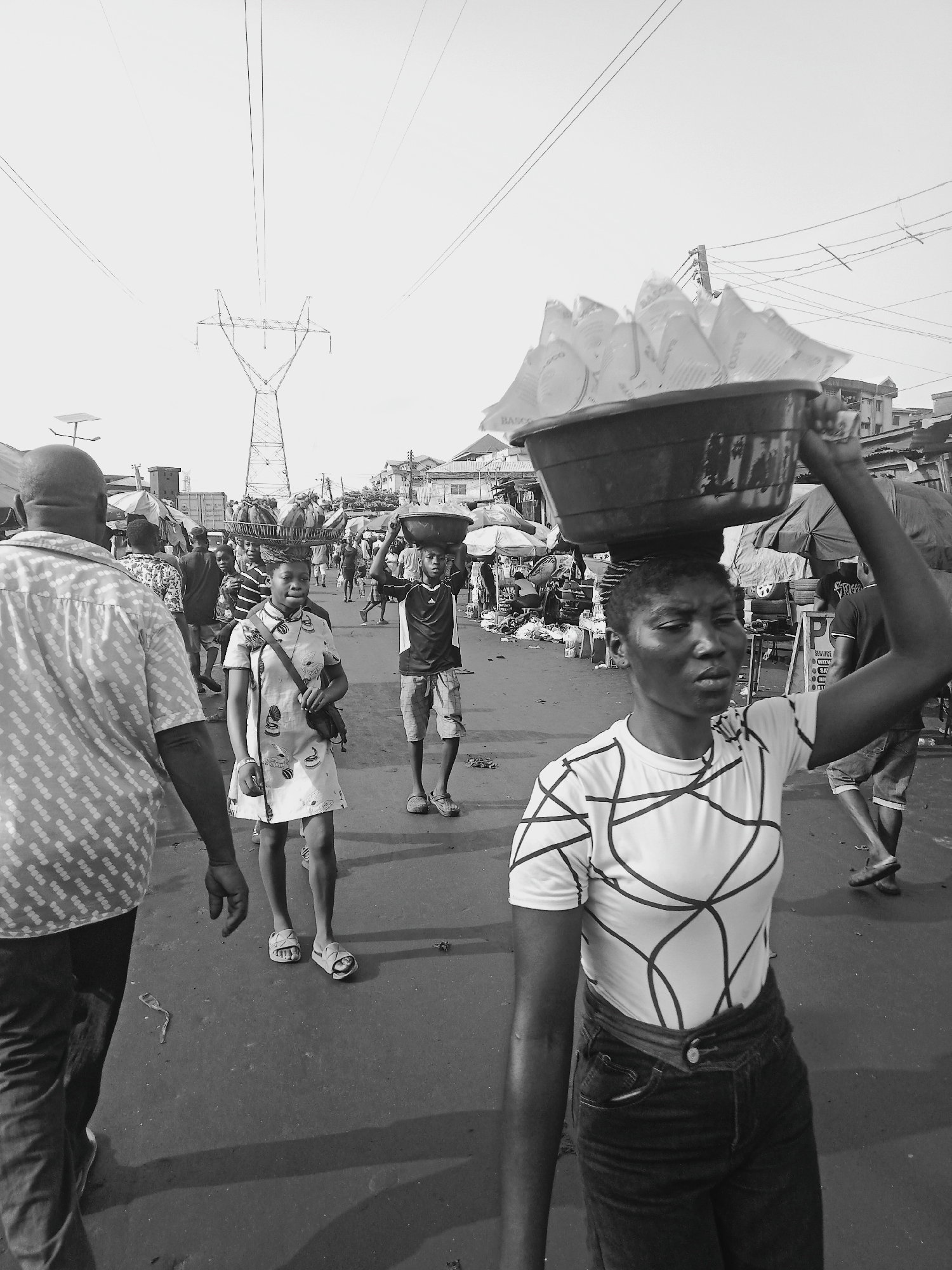 In Unknown, Unknown, a monochrome image captures a street bustling with people. Predominantly in the foreground, a woman carries a basin atop her head, her right arm raised for balance. She is dressed in a patterned shirt and dark jeans. Behind her and to the left, a young girl in a patterned dress walks with a purse slung over her shoulder. To her right, a boy carries a similar basin on his head, clad in a t-shirt and shorts. In the immediate foreground, a man in a patterned shirt walks away from the viewer. The background is crowded with people, stalls, and buildings. An electrical transmission tower stands in the distance, adding to the urban landscape.
