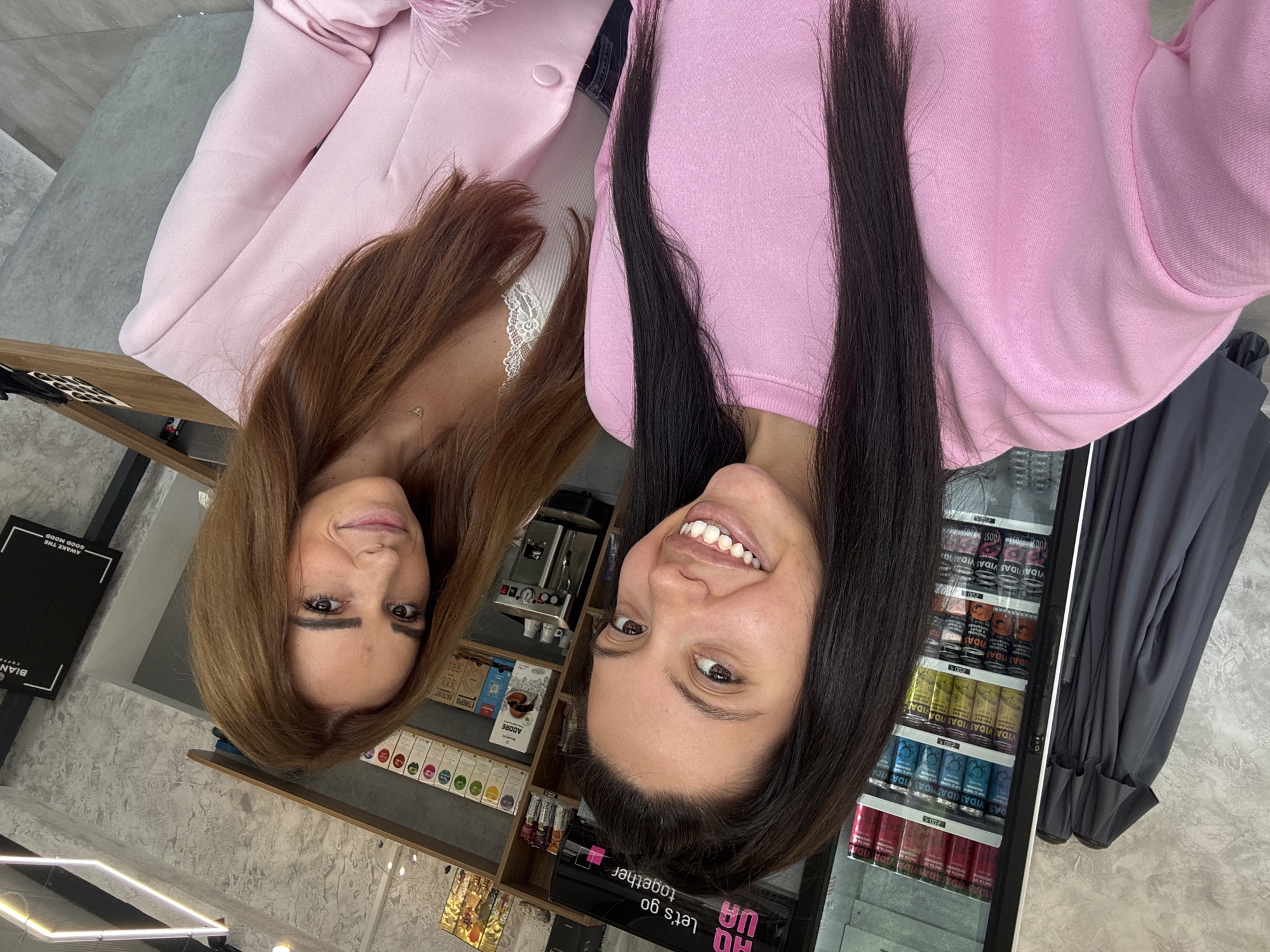 Two young women, one with long dark hair and wearing a pink sweatshirt, and the other with long auburn hair and wearing a white lace camisole and a pink blazer, are taking a selfie. They are standing in front of a cooler filled with beverages and shelves stocked with coffee and other products. The background shows a concrete-like wall and modern lighting fixtures. The scene appears to be inside a cafe or a shop in Sofia, Bulgaria, likely a modern establishment.