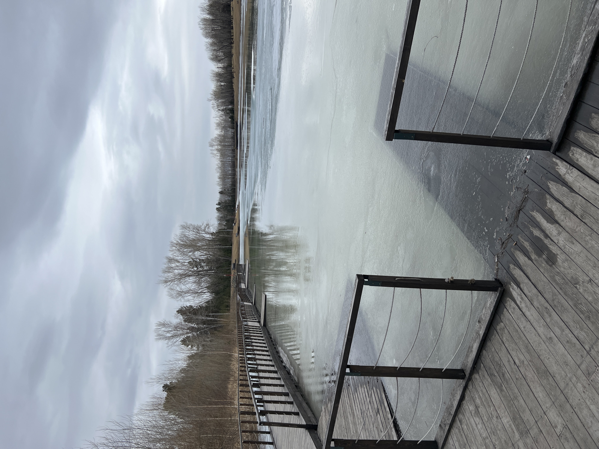 The image shows a frozen lake in Krasnoyarsk, Russia, seen from a wooden boardwalk. The lake is mostly covered with a thin layer of ice reflecting the overcast sky. Bare trees line the far side of the lake. The boardwalk features wooden planks and metal railings. The overall scene is bleak, depicting a cold, wintery day.