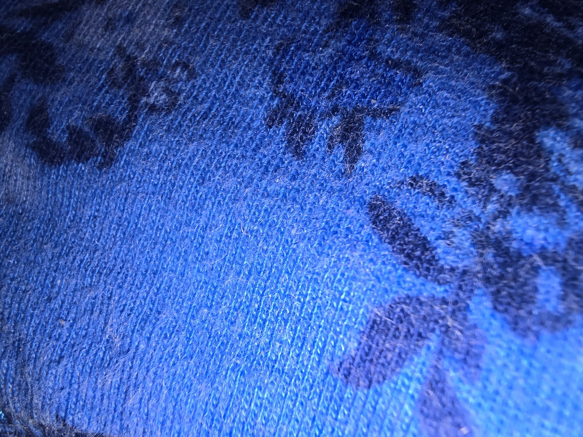 The image displays a close-up, macro shot of a textured blue fabric with a darker, abstract pattern. The fabric appears to be woven or knitted, with visible threads and a slight fuzziness, suggesting it could be a blanket, scarf, or upholstery. The lighting creates highlights and shadows, emphasizing the texture and depth of the material. The pattern is organic and irregular, with no discernible figures or symbols. There are no people, objects, actions, or interactions visible, and no environmental context is provided. Therefore, it is impossible to determine the location in Ulan-Ude, Russia, or any other specific location.