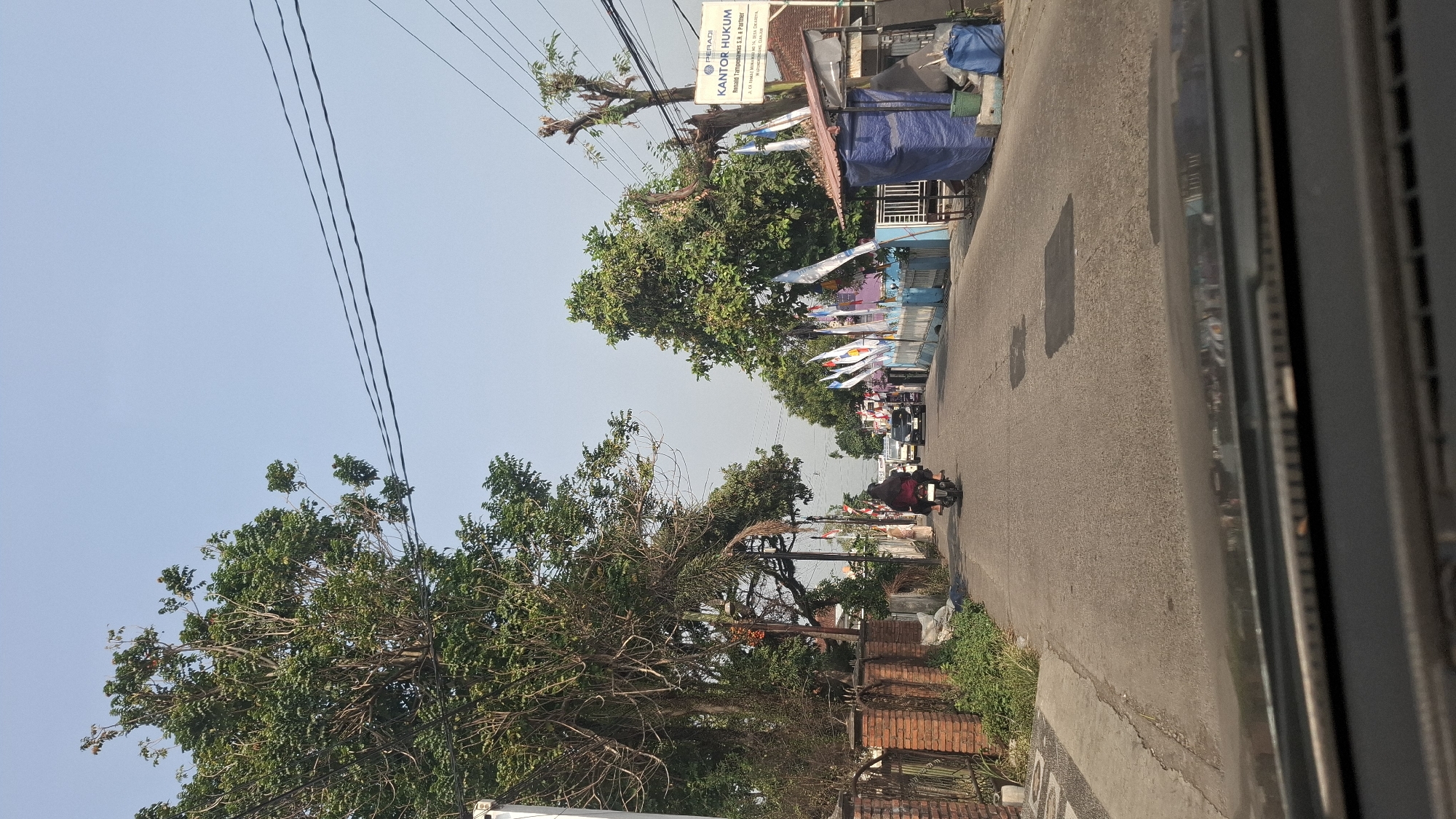The image shows a view from a car, looking down a residential street in Cikarang Utara, Indonesia. The street is paved and shows signs of wear with several dark patches indicating repairs. On the left side of the street, there is a brick wall bordering a property with lush green vegetation. A large tree with dense foliage stands prominently on the left, its branches reaching towards the power lines that crisscross the sky. Several other trees line the street, some with trimmed branches and others more wild.
A person on a motorcycle, wearing a dark jacket and pants, is traveling away from the camera down the center of the street. Further down the road, other vehicles, including a white truck and a dark car, are also visible. On the right side of the street, there is a blue fence decorated with numerous flags, most of which are white with blue lettering and logos, suggesting a local event or campaign. Beyond the fence, a small blue tarp-covered structure is visible, with blue bins next to it. A sign is mounted on a pole on the right side of the street, reading "PERADI KANTOR HUKUM" and providing contact information. The sky is clear and pale blue, indicating a bright, possibly sunny day. The overall impression is a typical urban or suburban street scene in Indonesia, with a mix of residential properties, vegetation, and infrastructure.