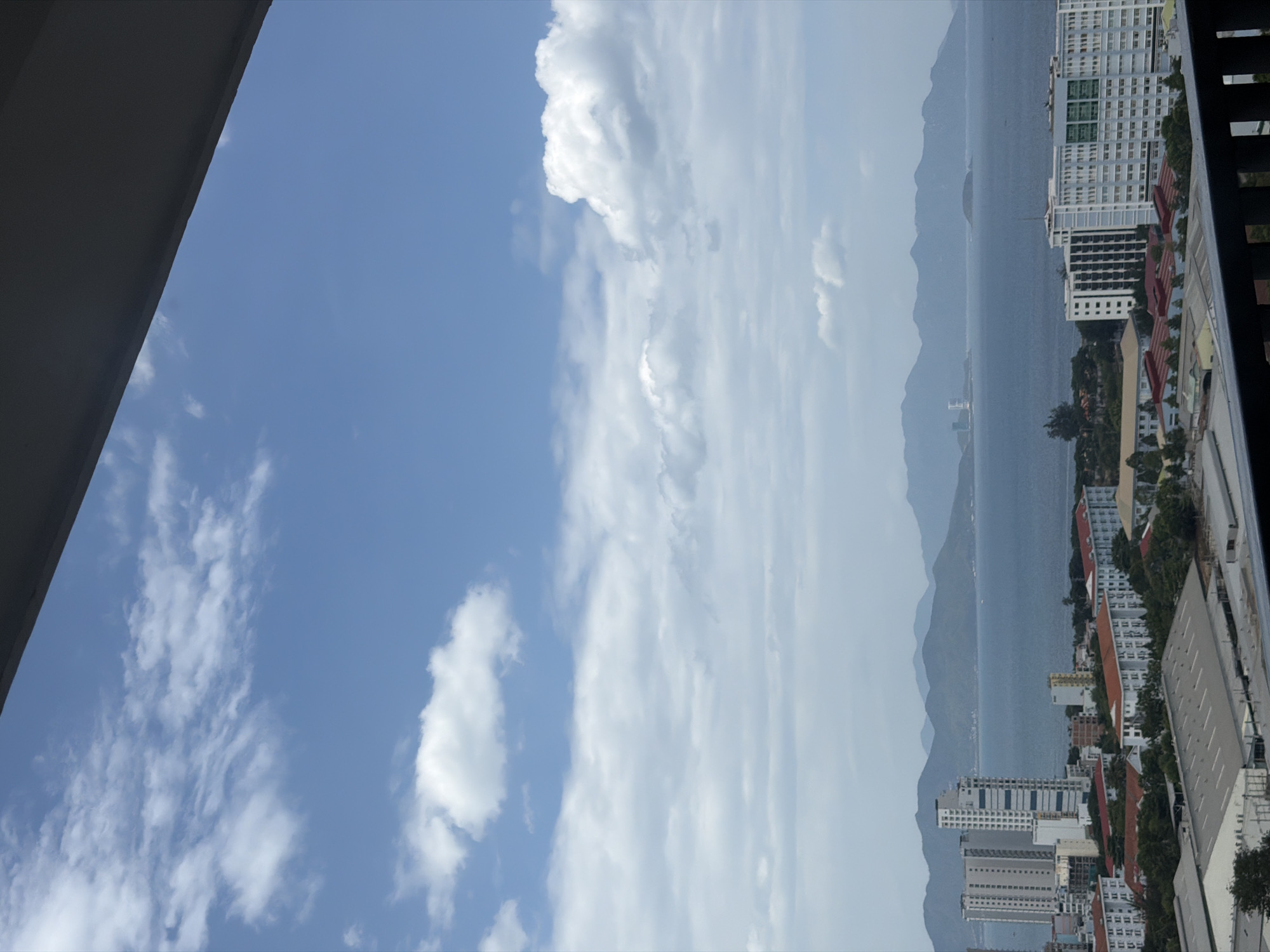 This is a high-angle, vertical, full shot of a city on the coast of Nha Trang, Vietnam. The sky is bright blue with scattered white clouds. In the foreground, a dark railing is visible, suggesting the photo was taken from a balcony or window. Below the railing, a cluster of buildings forms the cityscape. To the left, several tall, modern high-rise buildings with white facades and numerous windows stand out. Adjacent to these, a complex of lower-rise buildings with red tiled roofs stretches across the middle ground, interspersed with green trees. Further in the distance, a large body of water, likely the ocean, stretches to the horizon, where a range of blue-green mountains can be seen. The water is a calm, deep blue, with a few faint ripples. There are no people or vehicles clearly visible in the scene. The overall impression is a clear, sunny day over a coastal city.
