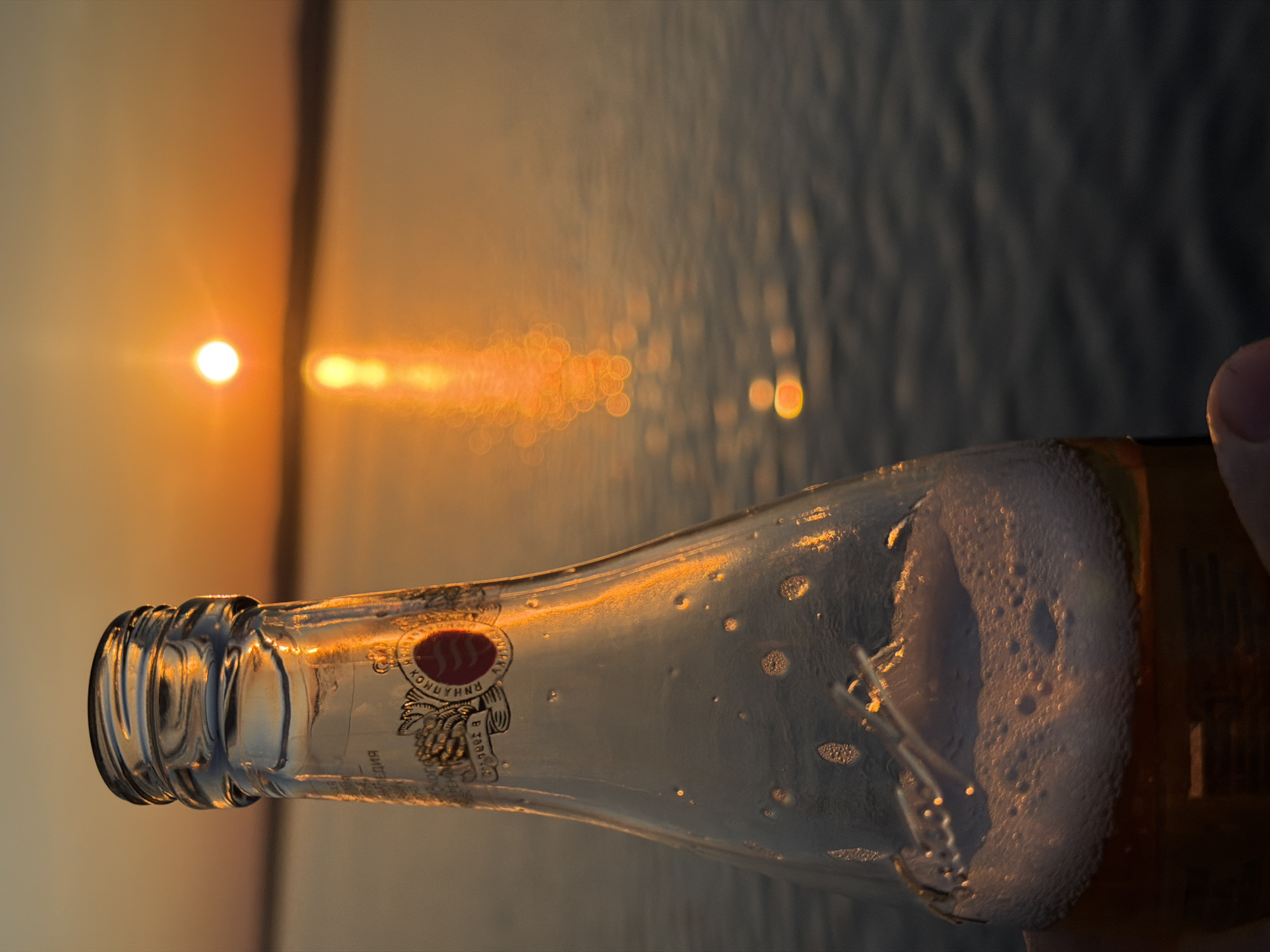 A horizontal medium shot focuses on a nearly empty, glass bottle of alcoholic beverage, likely beer based on the white foam, held by a person’s fingers visible on the right. The bottle is illuminated by the setting sun, which casts a bright reflection on the glass. The sun appears as a distinct point of light, with a series of blurry, circular reflections trailing towards the right on the water surface. The background is a blurred expanse of water and a soft, orange-tinted sky, indicating sunset or sunrise. The logo on the bottle is partially visible, featuring Cyrillic text and a symbolic design. The scene may be in the city of Chaikovsky, Russia, based on the Cyrillic text.