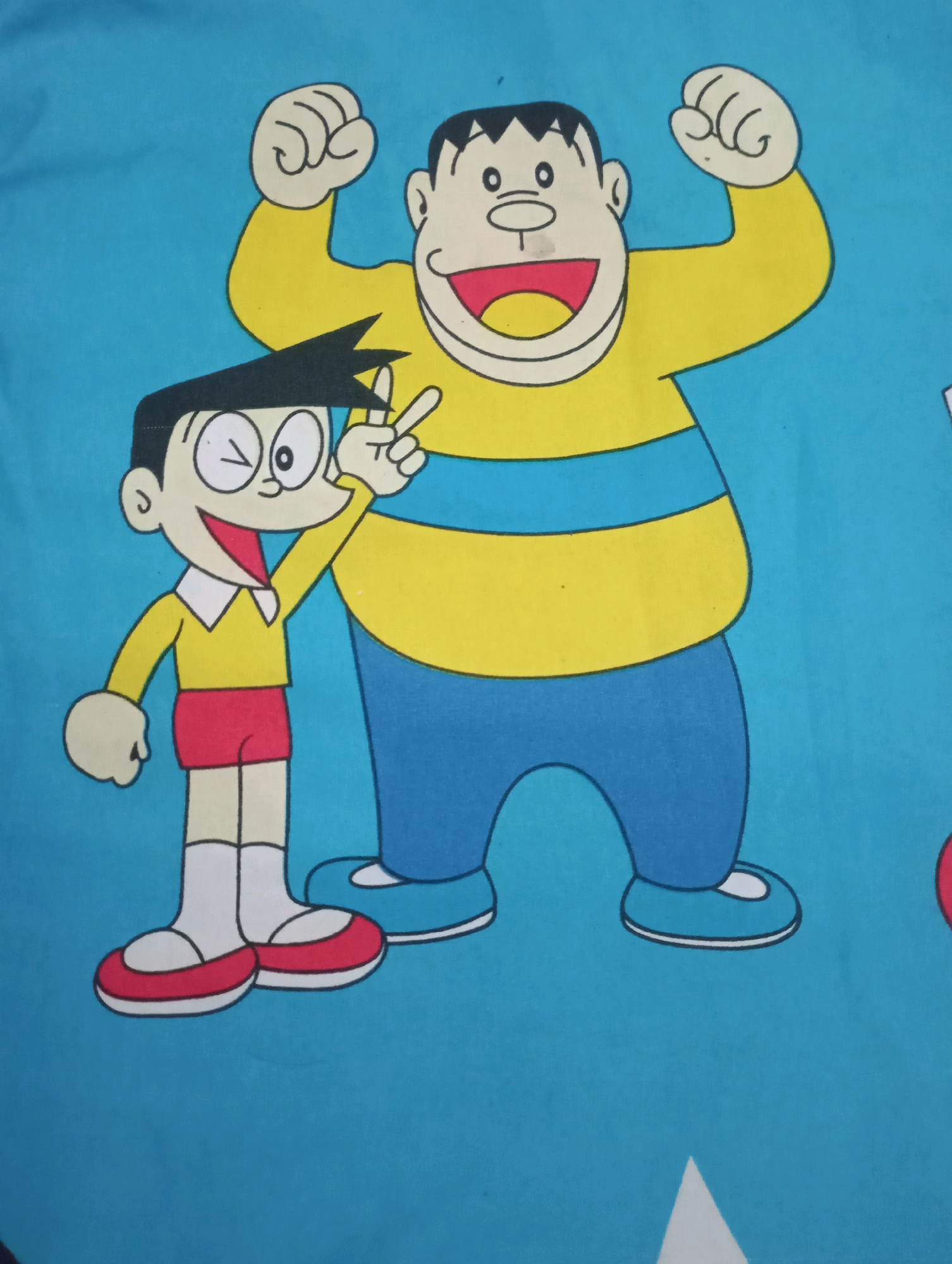 The image shows a cartoon depiction of two characters on a blue background. The character on the left is a young boy with black hair, wearing a yellow shirt with a white collar, red shorts, white socks, and red and white shoes. He is winking and holding up two fingers in a peace sign. The character on the right is a larger, older boy with black hair, wearing a yellow shirt with blue stripes and blue pants. He is flexing his arm. The cartoon appears to be printed on fabric, possibly a bedsheet, given the slight creasing visible. The image does not provide any information about a specific location in Kecamatan Haurgeulis, Indonesia.