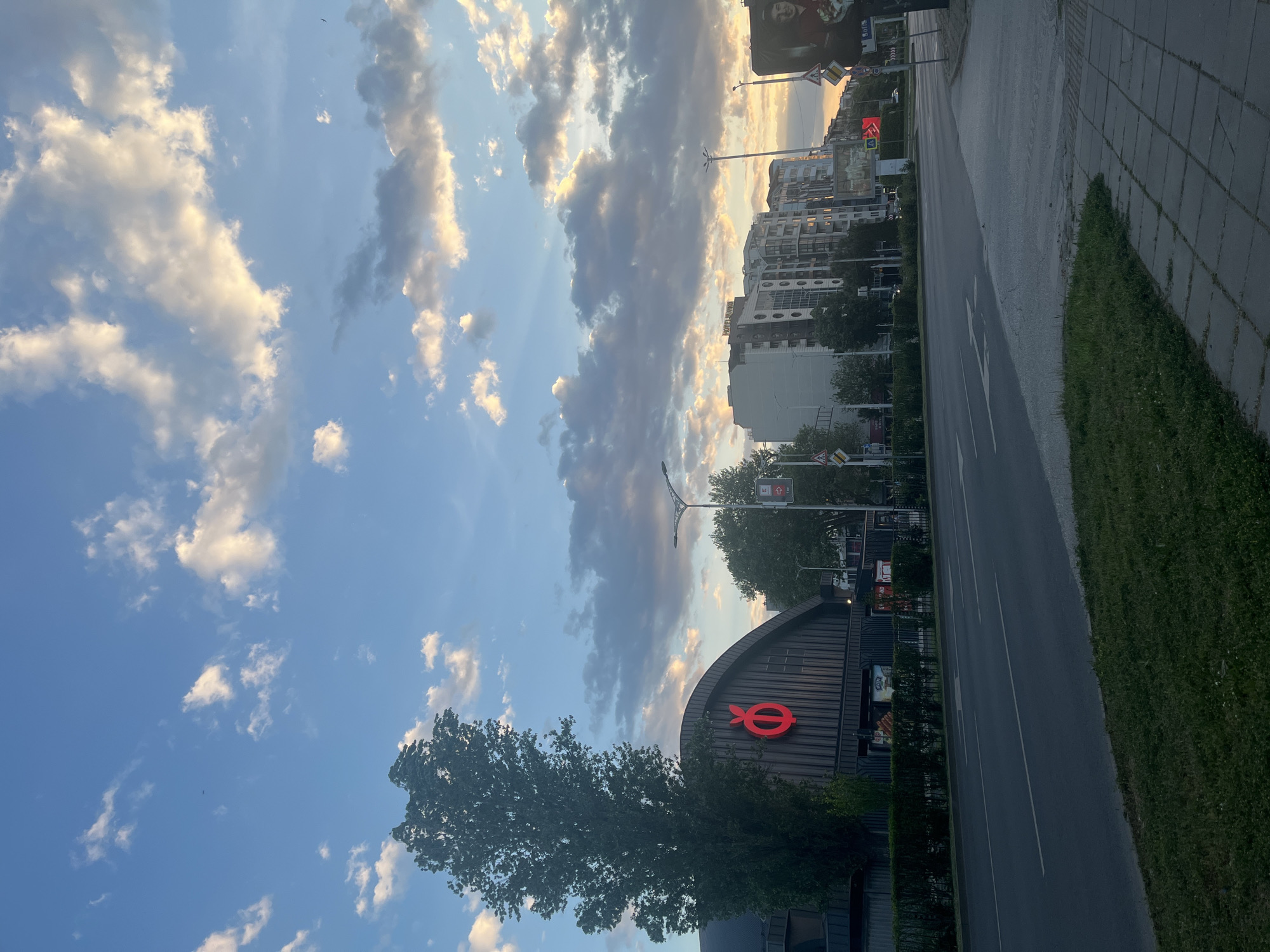 The image shows a street scene in Plovdiv, Bulgaria, with a large red "O" logo on a building visible in the foreground. The building's architecture is modern, with a curved roof. A mature tree partially obscures the building. Adjacent to the building is a road with directional arrows painted on it. A sidewalk lined with greenery runs parallel to the road. In the background, there are multiple buildings of varying heights, and the sky is filled with clouds, indicating either sunset or sunrise. Streetlights are present along the road.