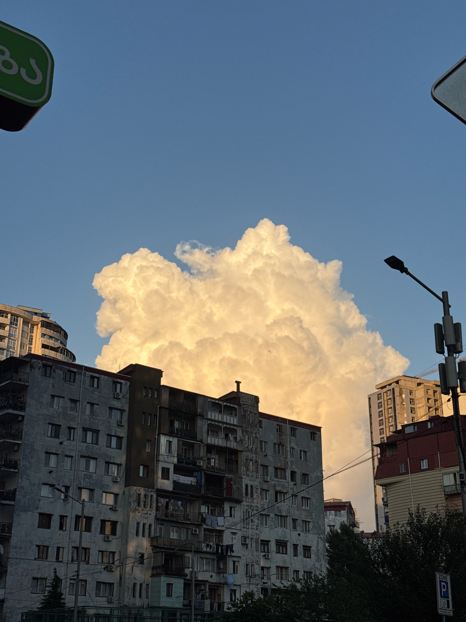 The image captures a cityscape in Batumi, Georgia, dominated by a massive, luminous cloud formation against a clear blue sky. In the foreground, a series of apartment buildings, some appearing weathered and aged, are visible. The architecture is characteristic of Soviet-era construction, with multiple stories and visible balconies. A streetlamp with attached cellular antennas stands to the right of the frame, also illuminated by the warm light. The bottom of the image shows a dark green canopy of trees and a parking zone sign. The overall impression is a blend of urban life and dramatic natural phenomena, with the golden light of either sunrise or sunset casting a glow on the scene.