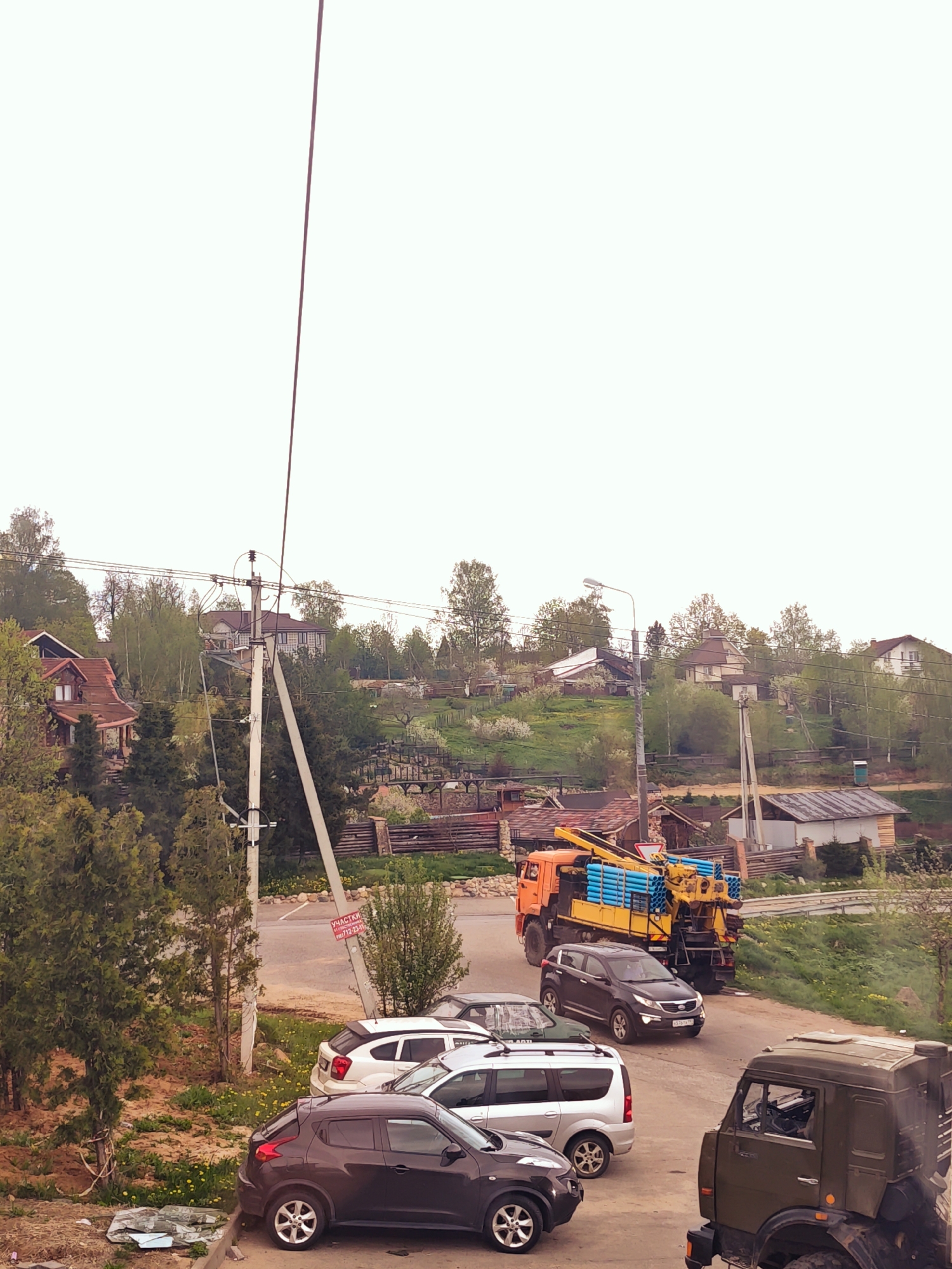 The photograph depicts a street scene in what appears to be the city of Ильинское, Russia, based on the Cyrillic lettering on a sign attached to a utility pole. The scene is a residential area with a mix of houses and greenery on a slight incline.
In the foreground, multiple cars are parked. These include a dark-colored Nissan Juke, a silver minivan, a white hatchback, and a dark SUV. Partially visible at the bottom right corner of the frame is a military-style truck painted in olive green.
Further down the street, a large orange truck is noticeable. It has a yellow platform carrying blue pipes. A dark SUV appears to be driving behind it.
In the background, the landscape includes houses of different architectural styles, scattered across the hill, interspersed with trees and other vegetation. Utility poles and power lines stretch across the scene.
The sky is overcast, suggesting the weather conditions may be cloudy.