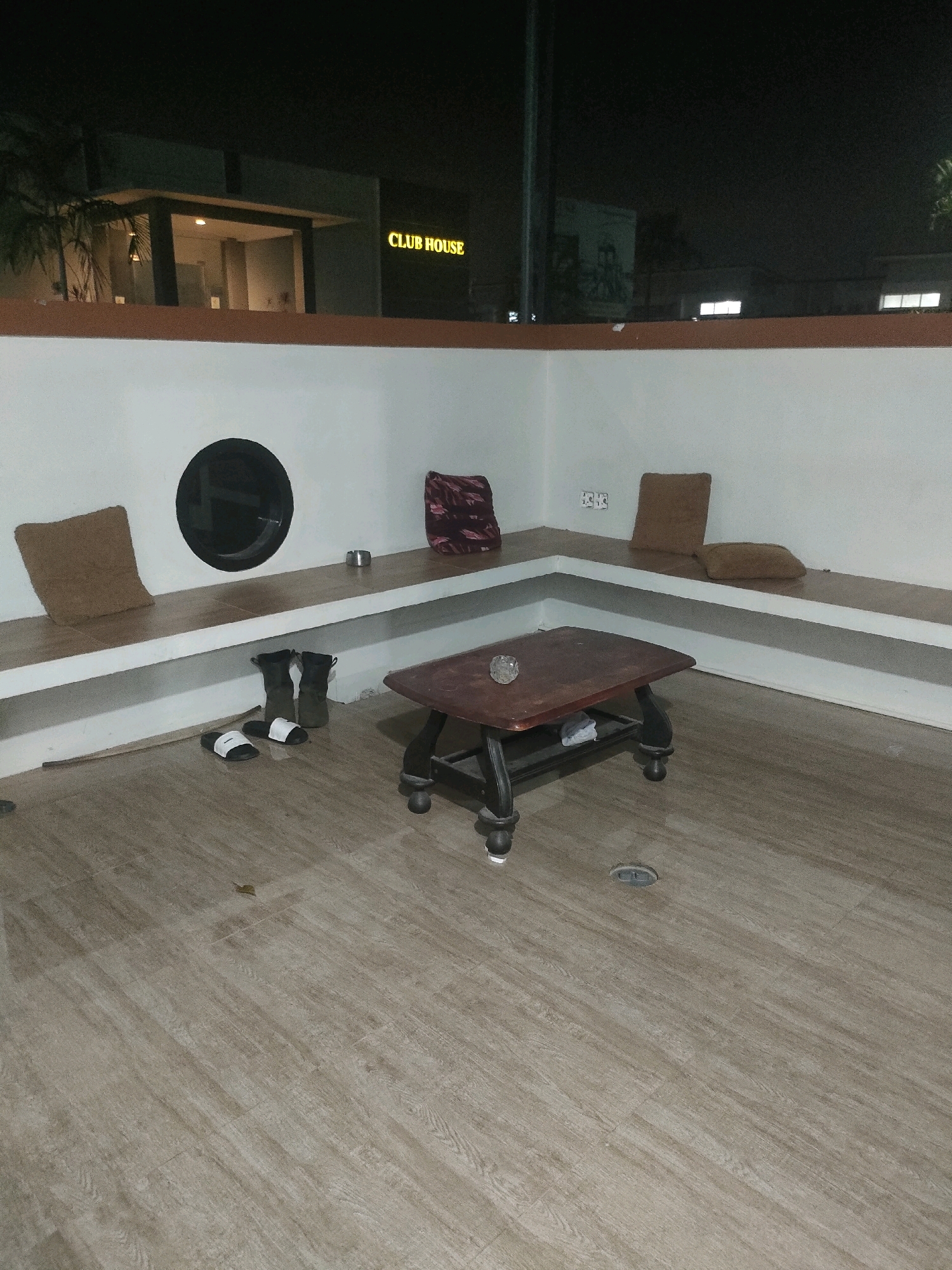 The image shows an outdoor seating area at night, likely part of a "CLUB HOUSE" as indicated by signage visible in the background. The seating area features a built-in, L-shaped bench made of light-colored wood or tile, with cushioned seating and a small, dark wood coffee table in the center of the indoor corner. On the bench, there are three cushions: one light brown, one darker brown, and one with a red and black abstract pattern. A small, metal ashtray sits on the bench. On the floor in front of the bench, there are a pair of black flip-flops and a pair of dark boots. The floor itself is tiled with a wood-like pattern. In the background, a building with illuminated windows and the "CLUB HOUSE" sign is visible. The overall scene suggests a relaxed, possibly communal outdoor space. The location in Batam, Indonesia, implies a tropical or subtropical climate, which is consistent with the outdoor setting.