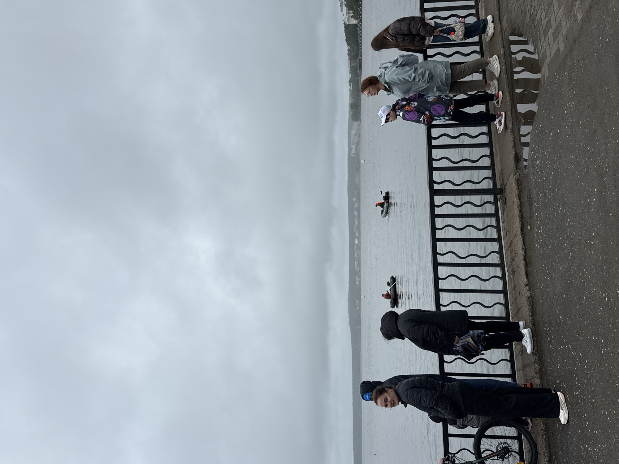A group of people are standing and walking along the embankment of Lake Votkinsk in Votkinsk, Russia. The sky is overcast, and the water is calm. A black metal fence separates the embankment from the lake. In the distance, two small boats are visible on the water, each with a person inside. On the embankment, a bicycle is parked to the left. A woman in a black puffer jacket and black pants is standing by the bicycle, facing left. Next to her, a person in a black hooded jacket is also facing left. Further to the right, a person in a black hooded jacket is standing, holding a small umbrella. Next to them, a young person with a white baseball cap is walking, wearing a colorful jacket with smiley faces. Behind them, a woman in a light green jacket and glasses is walking, and a young woman in a brown jacket is walking behind her. The ground on the embankment is paved with asphalt, and there are puddles of water on the ground. Buildings are visible on the opposite shore of the lake.