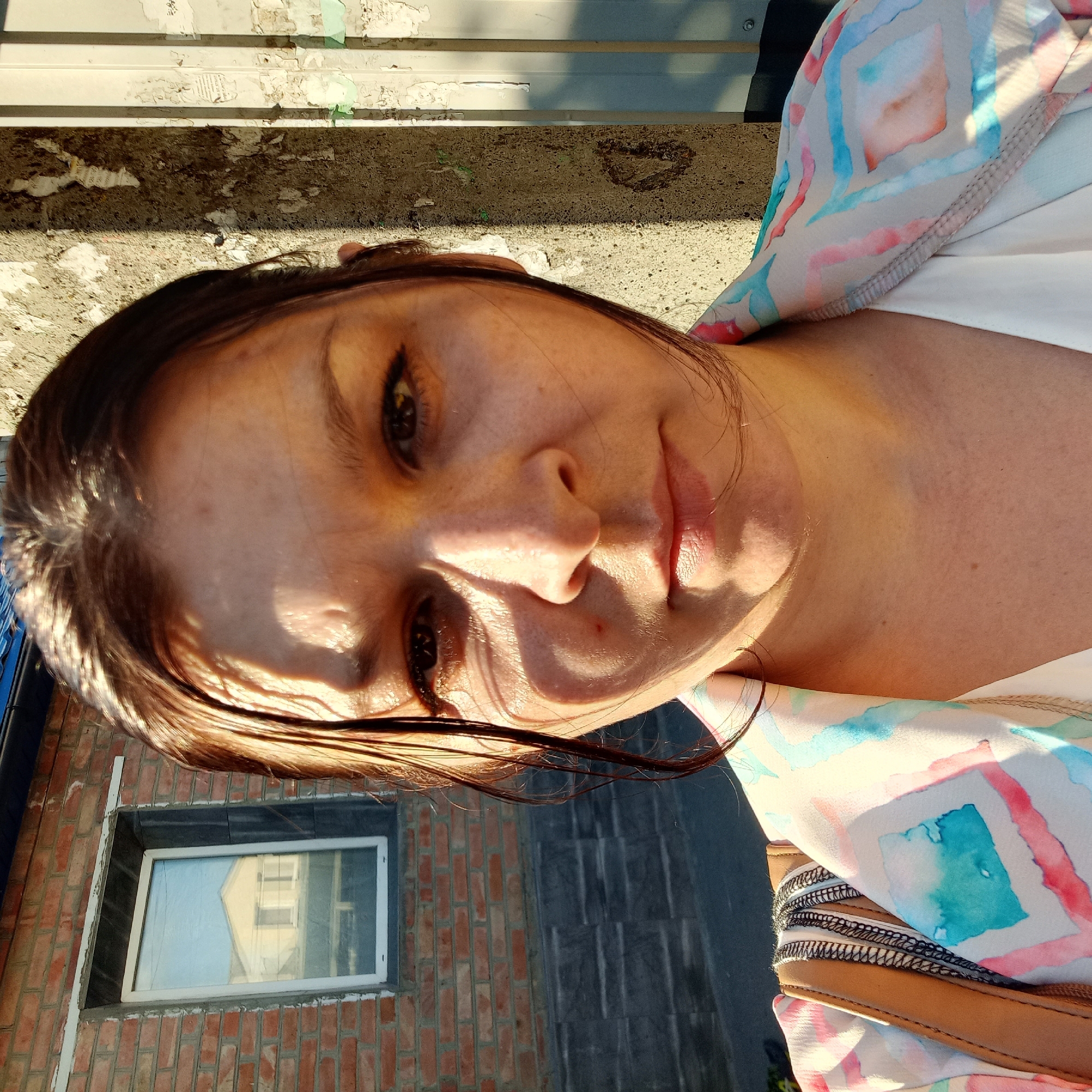 A medium shot selfie of a woman with wet, dark hair is captured outdoors, in Kropyvnytskyi, Ukraine. She is facing the camera directly, her expression neutral. Her face is partially illuminated by sunlight, creating shadows across her forehead and cheek. She is wearing a light-colored blouse with a patterned jacket over it, featuring abstract designs in pink, blue, and white. A brown leather bag is visible over her shoulder.
The background shows a brick building with a small, white-framed window, reflecting a yellow house. To the right, a textured concrete pillar with peeling posters and some vertical, off-white corrugated metal panels form the backdrop. The ground in the lower portion of the image appears to be asphalt. The scene suggests an urban or semi-urban outdoor environment.