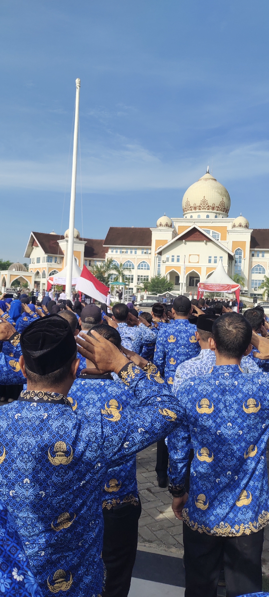 The image captures a scene in Meunasahaluedrienlhoksukon Barat, Indonesia, with a large group of people standing at attention, saluting toward a flagpole. The individuals are wearing similar blue shirts adorned with a repeating emblem. A red and white Indonesian flag is visible near the flagpole. In the background stands a substantial building with a distinctive dome structure. The sky is clear and blue, suggesting a sunny day.