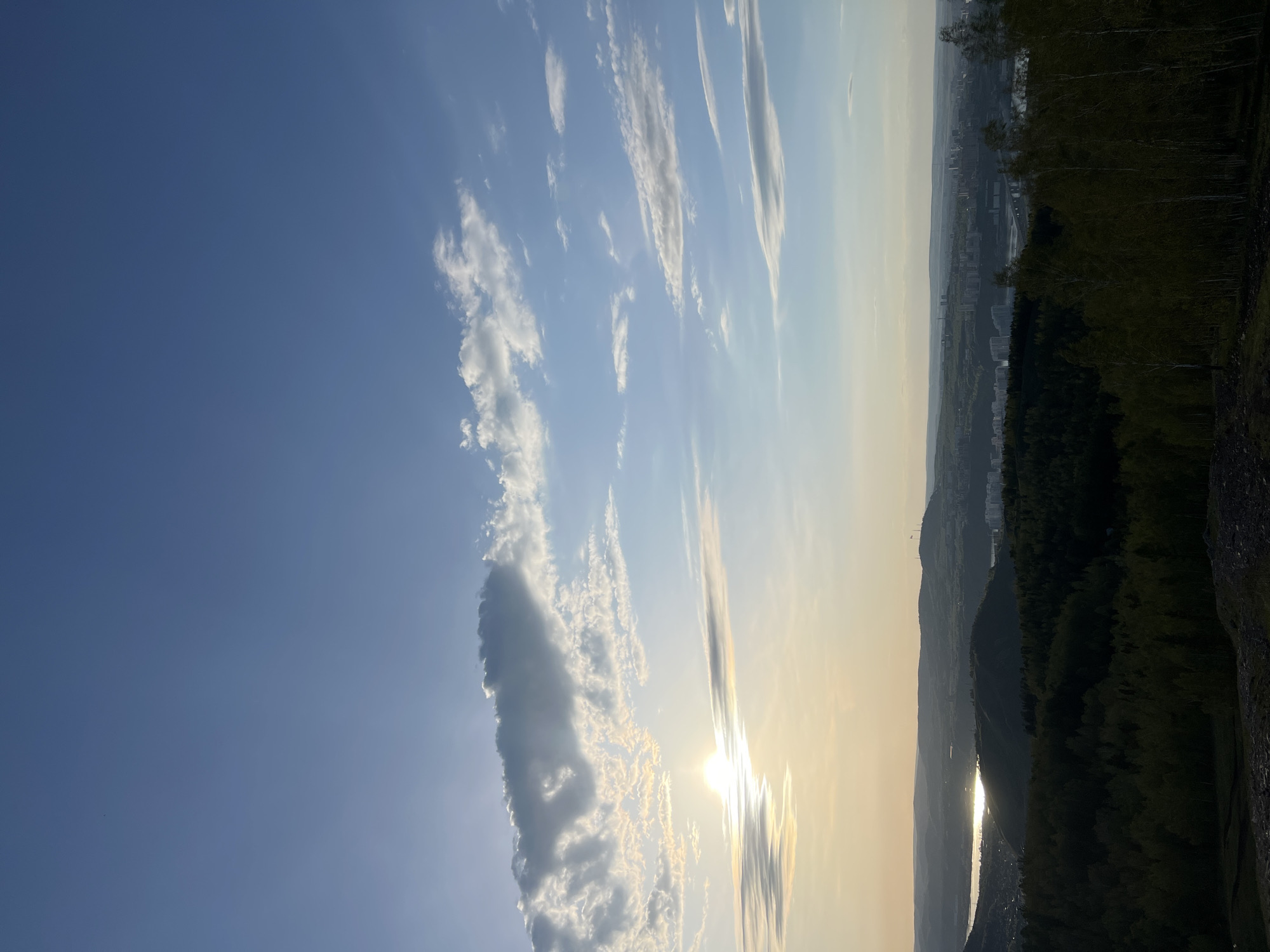 The image captures a landscape at sunset, possibly in Krasnoyarsk, Russia, given the information. The sky dominates the left portion of the frame, featuring a clear blue gradient with scattered cumulus clouds and a column of larger clouds. The sun is obscured by clouds, casting a golden light across the sky and a shimmering reflection on what appears to be a river or body of water below.
The right side of the image includes a wooded area and glimpses of urban structures, suggesting a city skyline. There is a dark edge in the bottom right corner which appears to be a hillside. The image has a slightly hazy quality.