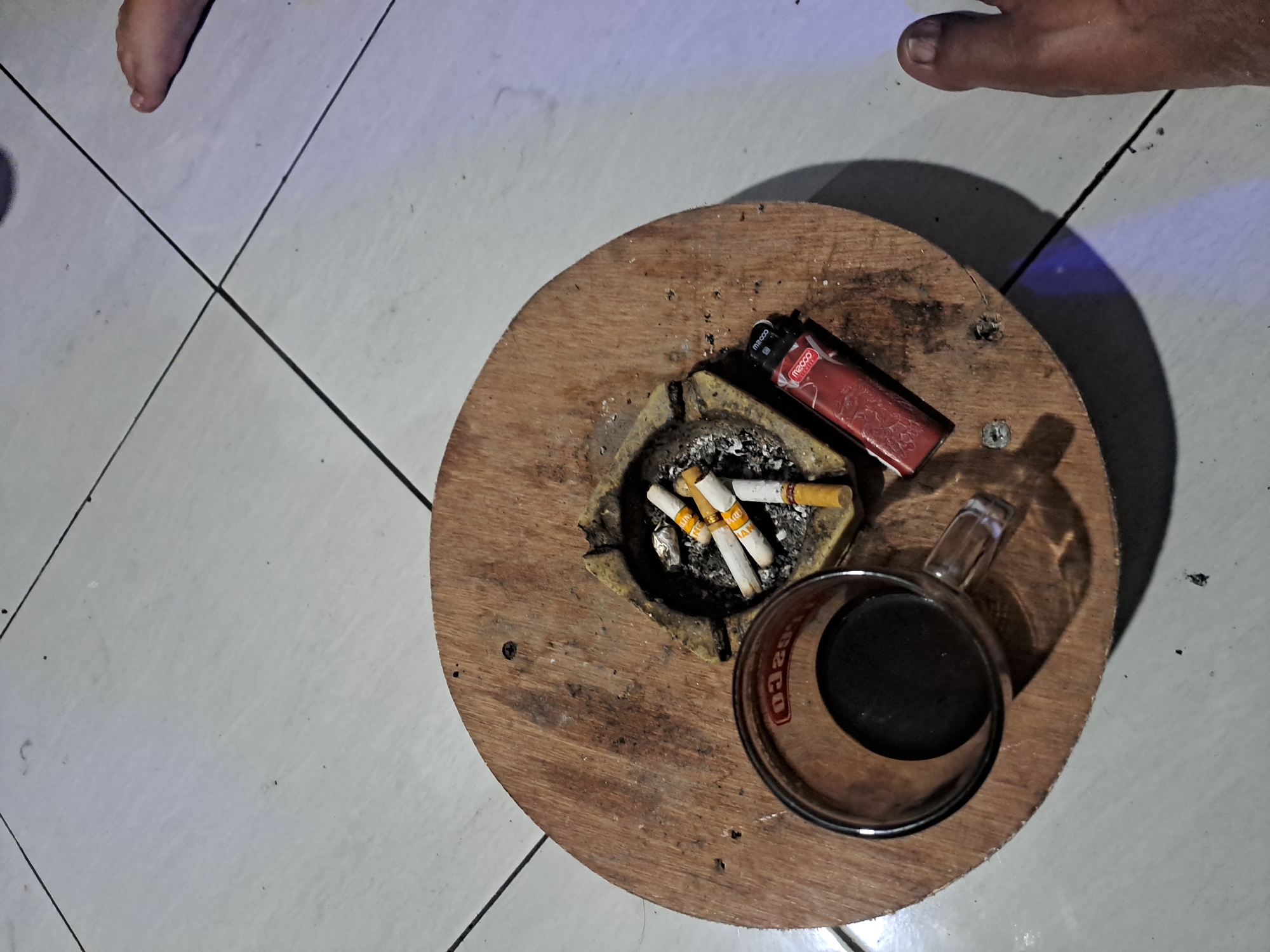 A close-up, overhead shot captures a wooden board with a glass of dark liquid, an ashtray with several cigarettes, and a red lighter. The subjects are placed on a tiled floor in what appears to be an indoor setting. The lighting suggests it might be evening or a dimly lit room. The overall atmosphere is one of casual relaxation, possibly in a home or a small establishment. The scene is devoid of any explicit indication of the city of Kebonagung, Indonesia, as no identifying landmarks or signs are visible.