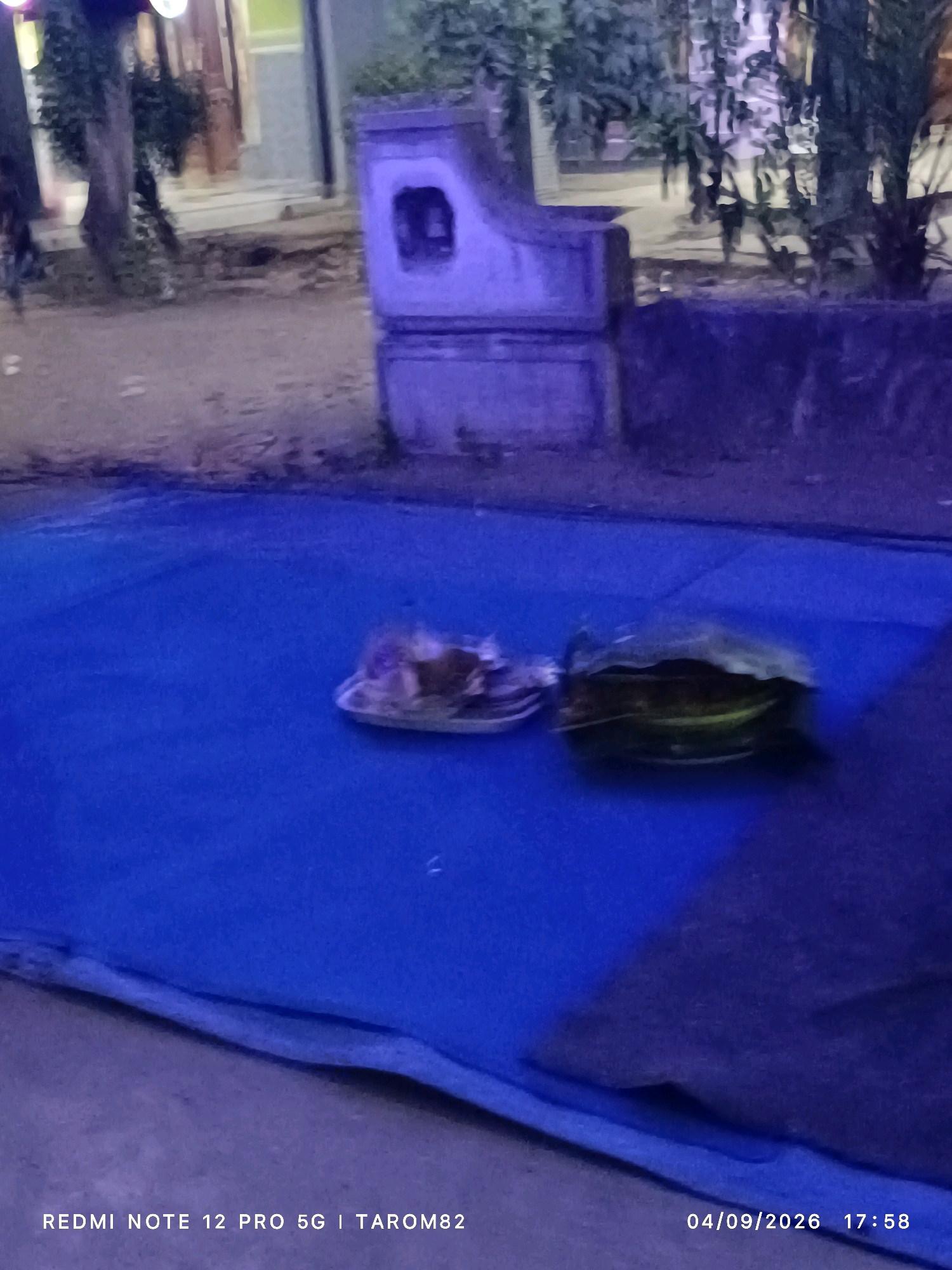 In Rejoagung, Indonesia, a blue tarp lies on the ground, possibly as a makeshift mat or covering. On the tarp, two plates hold offerings or food items. One plate contains a pile of what appears to be food, possibly rice and other dishes, while the other holds a large leaf or bowl with a dark filling. In the background, a building with a pale facade and a dark entrance stands, flanked by trees. A stone structure, possibly part of a bench or a decorative element, is visible to the left of the building. The scene is dimly lit, with the blue tarp dominating the foreground. The timestamp on the image indicates the date as April 9, 2026, at 17:58.