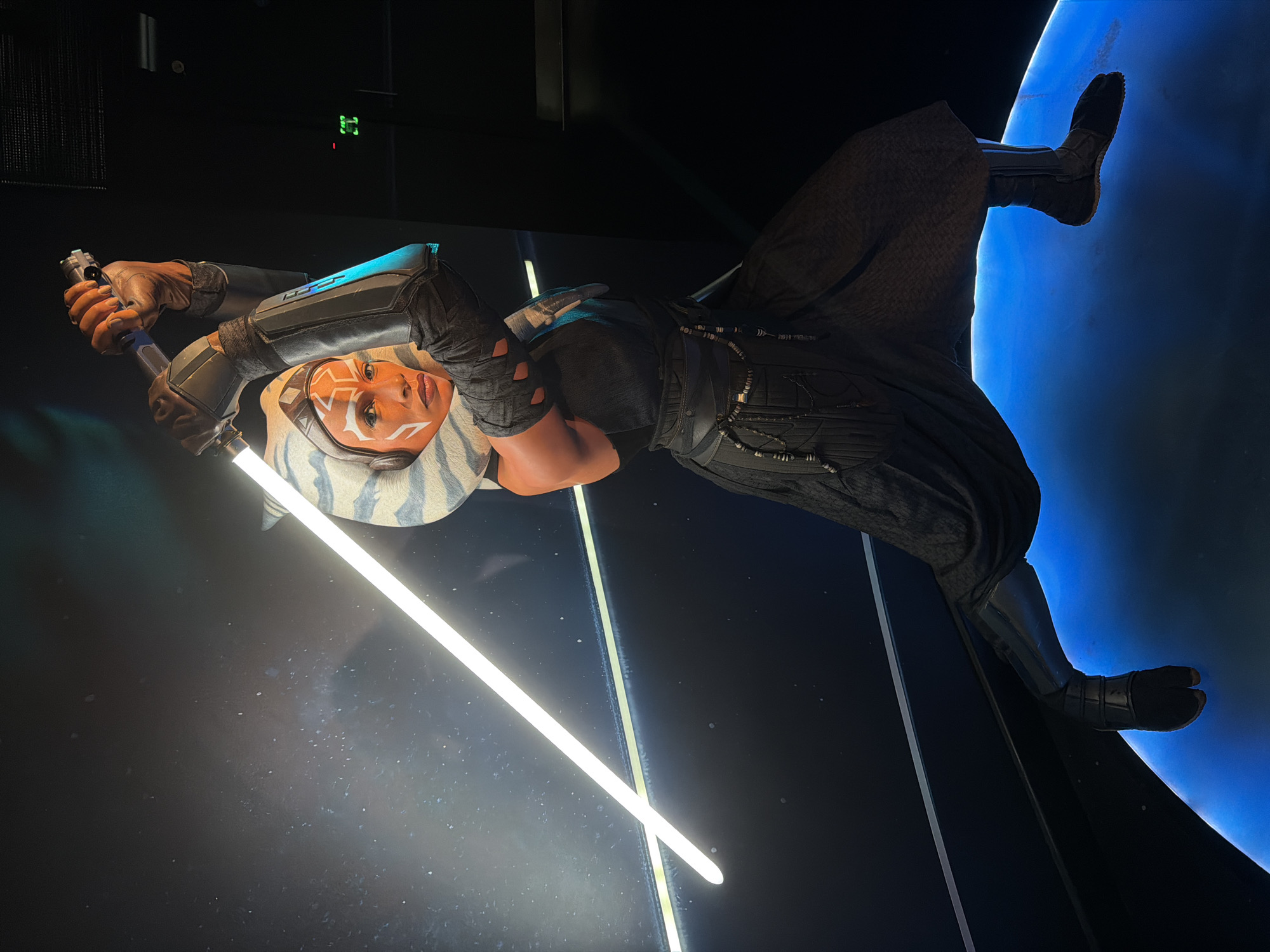 The image shows a statue of Ahsoka Tano, a character from the Star Wars franchise, holding two lightsabers. She is wearing a dark outfit with light blue accents on her arm guards and head tails. The statue is standing on what appears to be a blue planet-like structure. The background is dark, with a faint green light fixture visible in the upper left corner. The image could be from a museum exhibit or a promotional display. There is no information in the image about the city of London, United Kingdom.