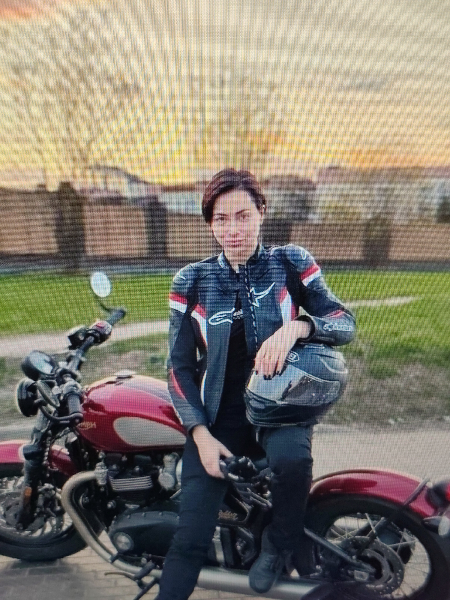 A young woman with short dark hair, wearing a black and white motorcycle jacket with red accents, black pants, and black boots, sits on a red motorcycle. She holds a black helmet in her right hand, resting it on her lap. Her left hand is placed on the motorcycle's gas tank. The motorcycle is parked on a paved surface, with a grassy area and a fence in the background. The sky is a hazy orange and yellow, suggesting either sunrise or sunset. The scene is likely outdoors, possibly in a residential area or a park, given the presence of a fence and grass. The overall impression is one of a casual pose with a motorcycle. The location is identified as Syktyvkar, Russia.