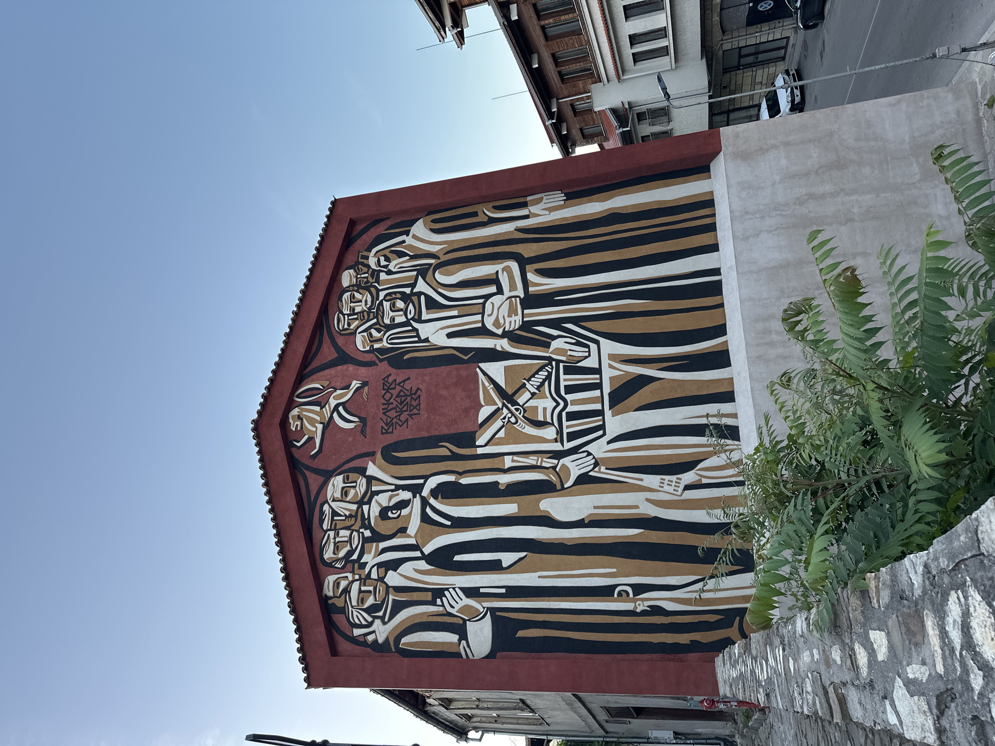 The image shows a mural on the side of a building in Veliko Tarnovo, Bulgaria. The mural depicts a group of men in traditional clothing standing around a table with a book and a weapon on it. The mural also features a lion emblem and text that reads "REHOVA ZAKARA 1835". The building is painted red and has a tiled roof. In front of the building, there is a stone wall and some greenery. The street in front of the building is paved with cobblestones and there are other buildings visible in the background. The sky is clear and blue.