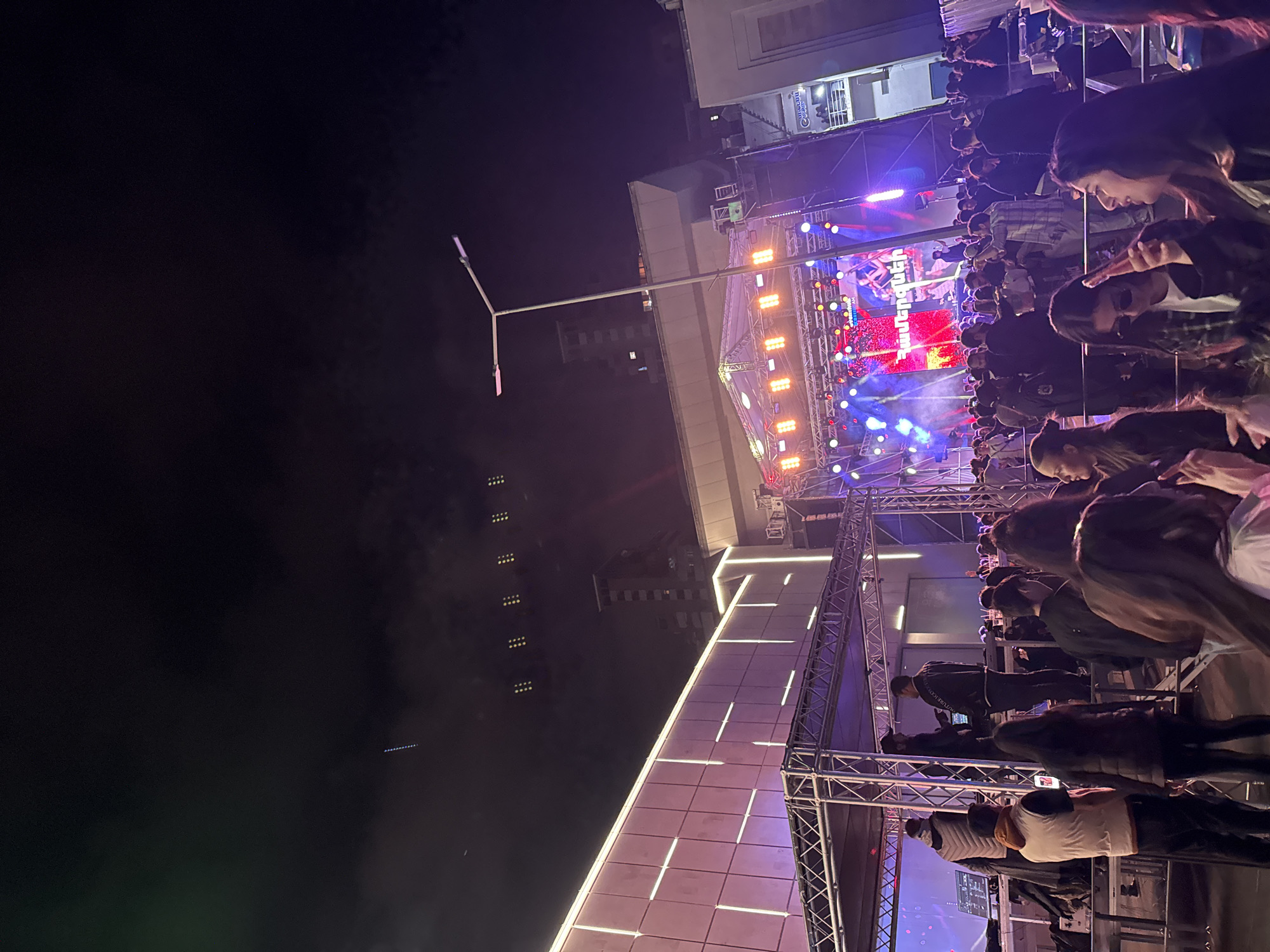 This image depicts a night scene at a concert or event in Yerevan, Armenia. A stage with bright lights and a large screen displaying an image is visible, with a crowd of people in the foreground watching. The people appear to be young adults. To the left of the stage is a streetlight and the dark sky. The perspective suggests the photo was taken from an elevated position, perhaps a balcony or a raised platform. The overall environment is urban and lively.